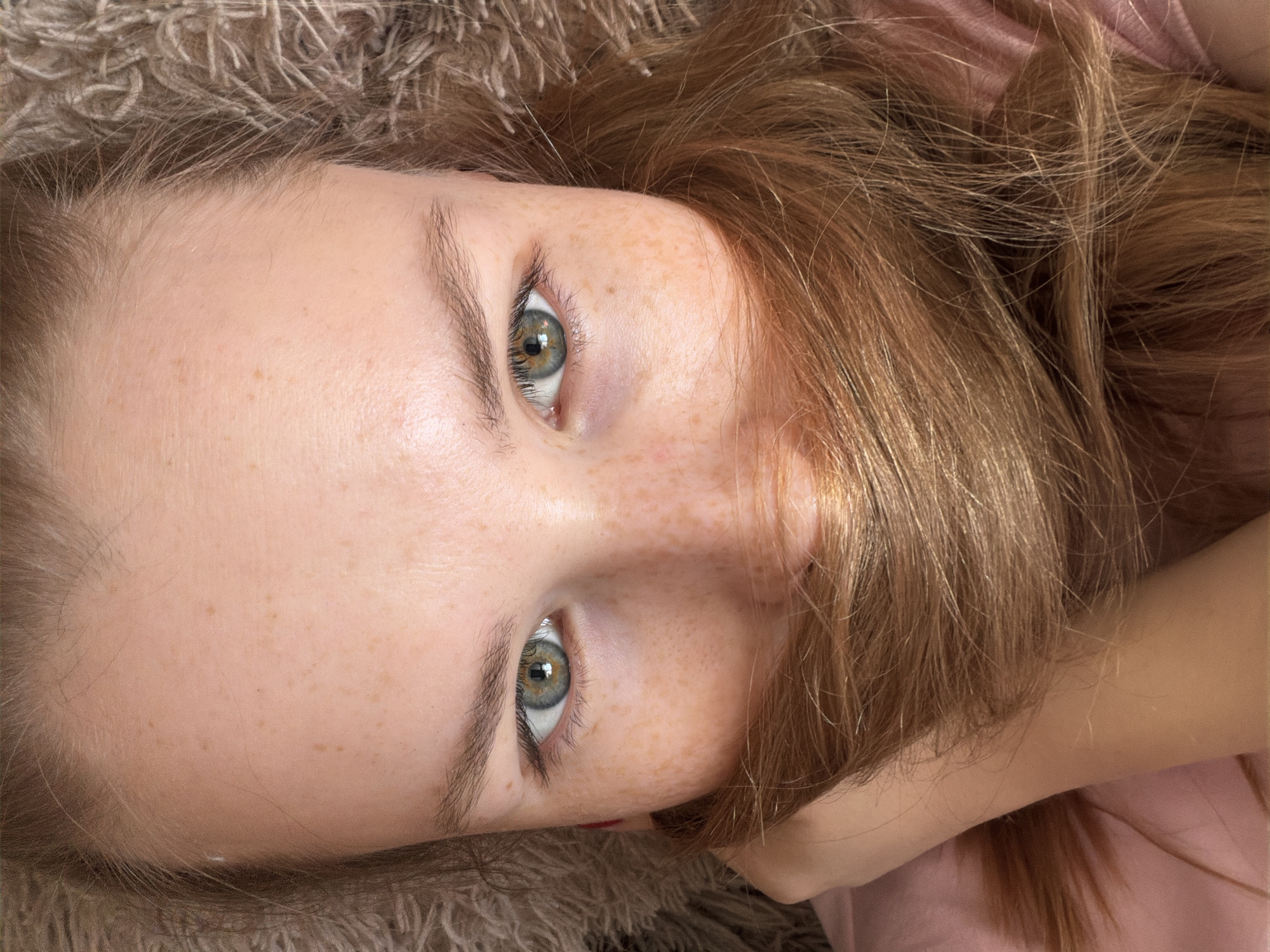 A close-up, high-angle shot shows the face of a fair-skinned person with light brown hair partially obscuring the lower right portion of the face. The person has freckles, light eyebrows, and eyes that appear green with a central dark ring. The background includes a beige, textured material, possibly a carpet or blanket, in the upper left corner. The person seems to be wearing a light pink garment. No specific location or action can be determined from the visual content of the image alone.