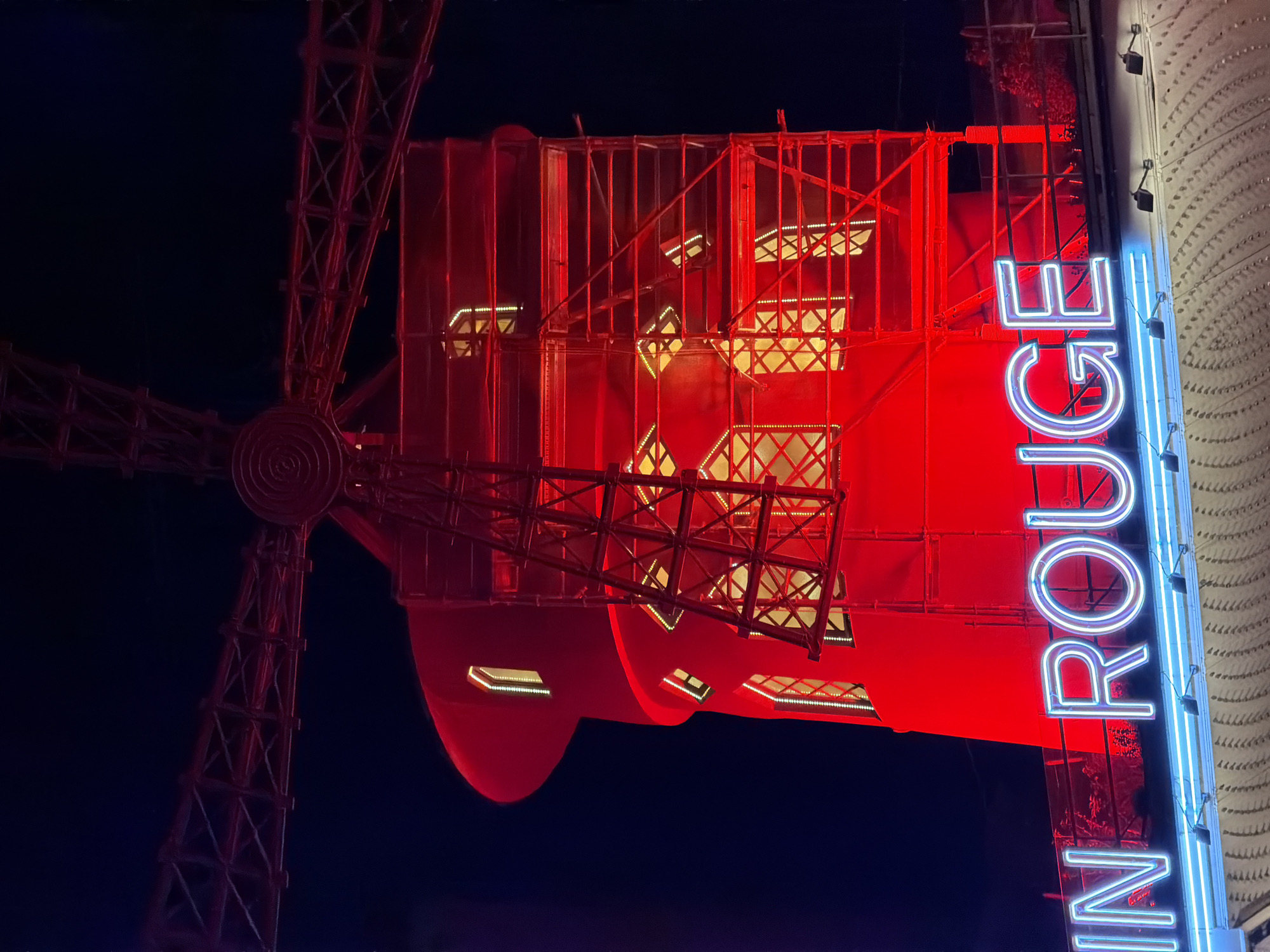 The image shows a nighttime view of the Moulin Rouge in Paris, France. The windmill is the focal point, illuminated in red. A blue and white neon sign reading "IN ROUGE" is positioned vertically on the building. The sky is dark, and the overall lighting is dramatic, emphasizing the Moulin Rouge's iconic presence.