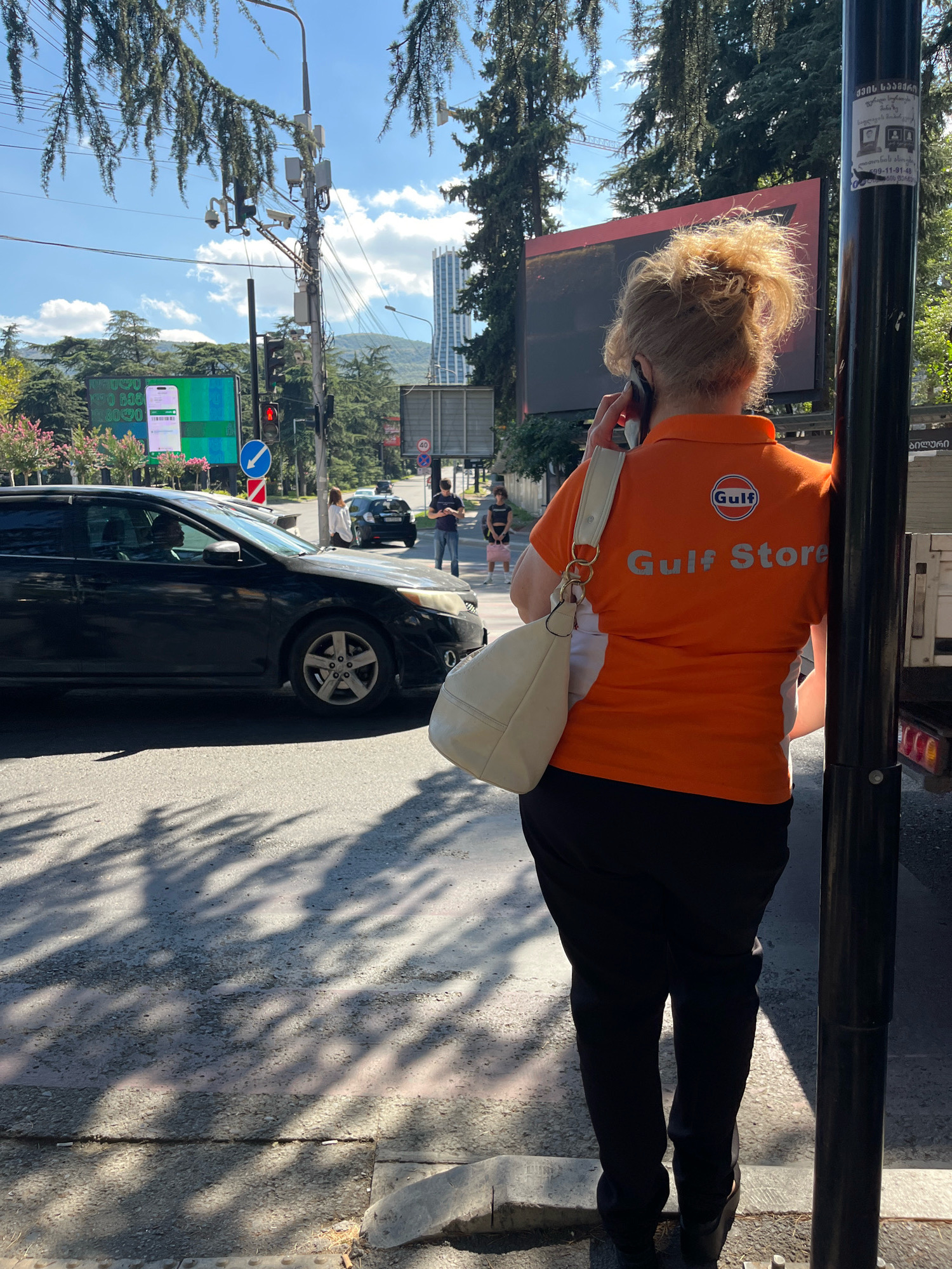 A woman with blonde hair pulled back is standing on a sidewalk next to a black utility pole and is speaking on her phone. She is wearing an orange polo shirt with a white collar and a white stripe down the side, and a white shoulder bag. The shirt has "Gulf" and "Gulf Store" printed on the back in silver lettering. She is wearing black pants. In the background, to the left, a black car is driving on the street. Further in the background, there are trees, traffic lights, a large billboard displaying a green and white advertisement with a phone screen, and a tall building. To the right, there is a white truck with a red stripe. The scene appears to be a street in Tbilisi, Georgia.