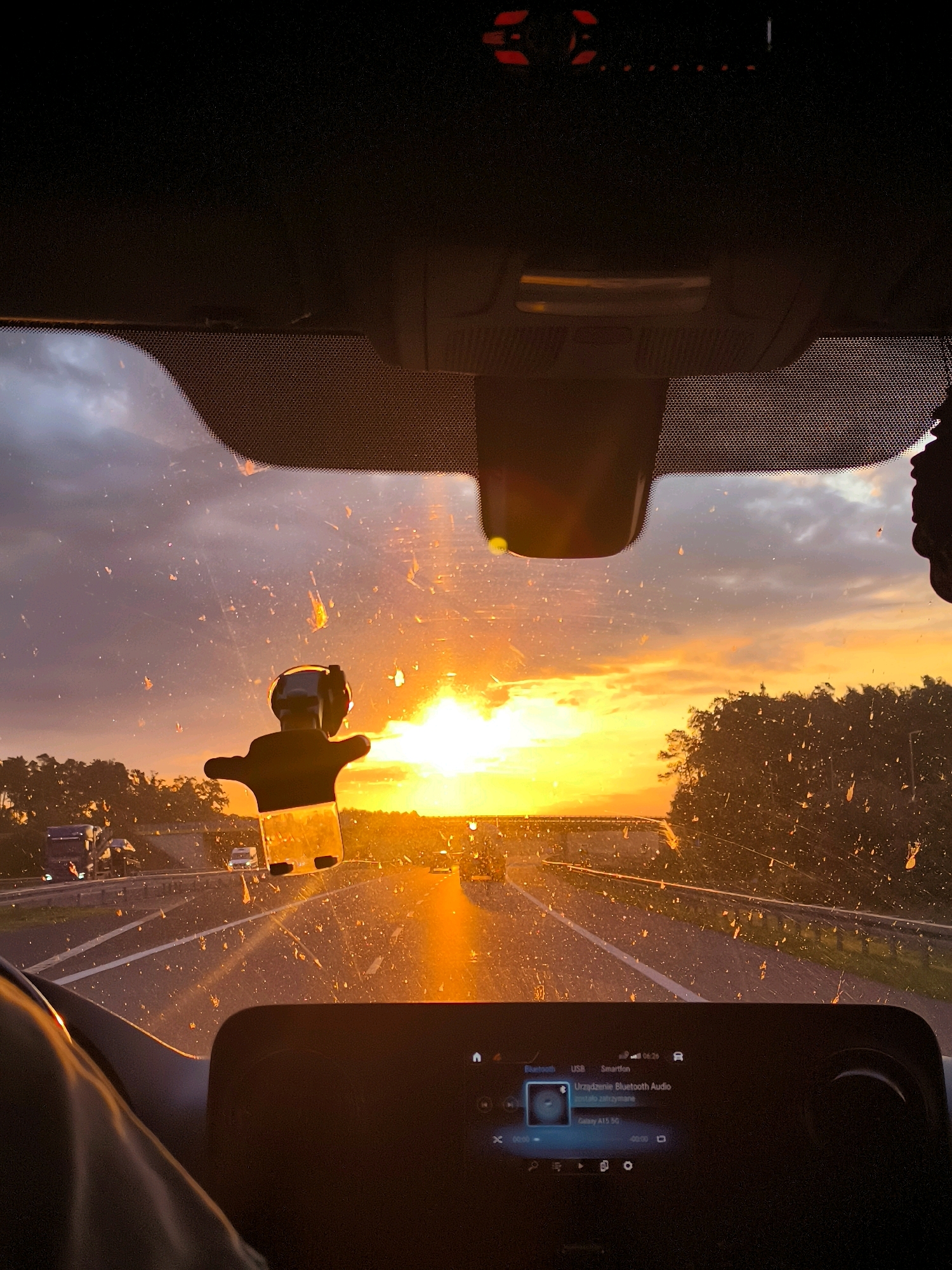 The image shows a view from inside a vehicle traveling on a highway at sunset. The sun is low on the horizon, casting a warm, orange glow across the sky and the road. The windshield is dirty with numerous spots and streaks, obscuring the view slightly. In the foreground, a dark dashboard with a digital screen is visible, displaying media information. A black car accessory, possibly a phone mount, is attached to the windshield. The highway is bordered by trees and other vehicles, including trucks and cars, are seen in the distance. The scene suggests a journey in Olesin, Poland, during a beautiful sunset.
