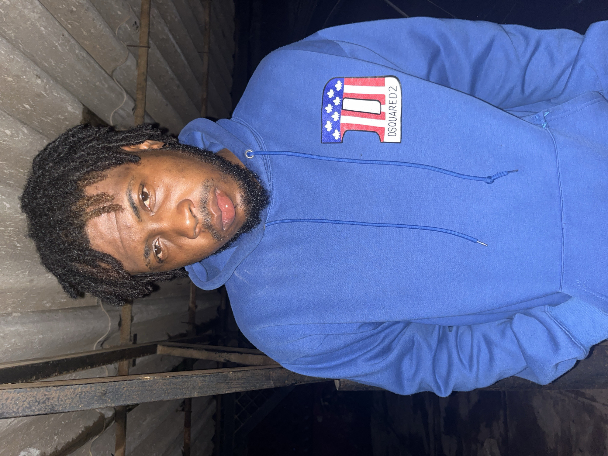 A medium shot captures a Black male standing in what appears to be an outdoor, possibly urban setting. He is wearing a blue hooded sweatshirt with a red, white, and blue patch on the left breast featuring the text "DSQUARED2". The background shows corrugated roofing material and some exposed wooden beams or supports, suggesting an unfinished or utilitarian structure. There are no clear indications of specific city landmarks, making it difficult to pinpoint the exact location within Jos, Nigeria. The lighting is dim, possibly indicating nighttime or an indoor setting with limited light. The individual's expression is neutral.