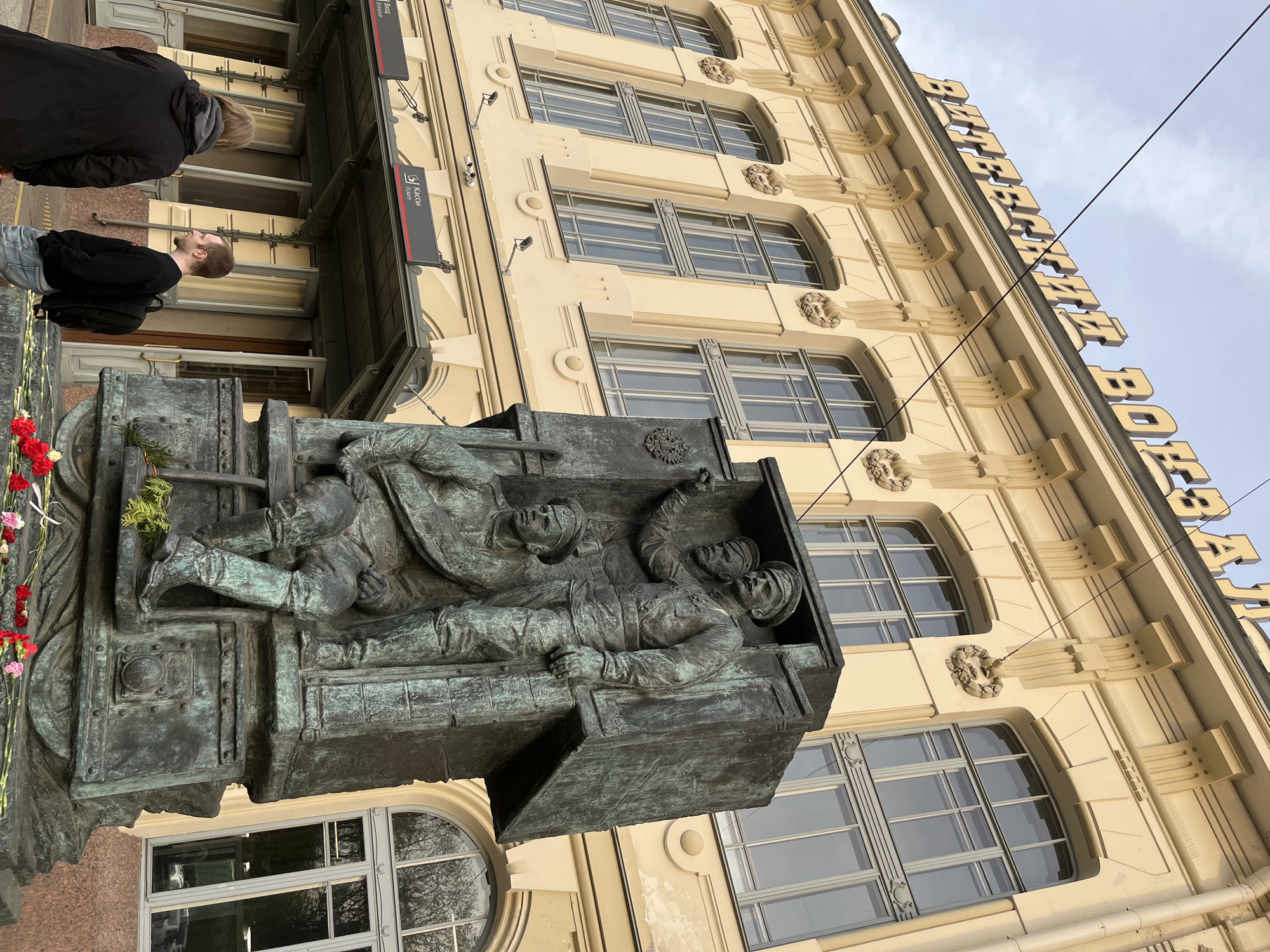 The image captures a street scene in an unknown city. The focal point is a bronze-colored sculpture depicting three figures lying down, seemingly inside a train car. The sculpture is mounted on the exterior of a multi-story building with a cream-colored facade and multiple windows. The building's architecture includes ornate details. A sign with large block letters is visible atop the building. To the left of the sculpture, two people are standing and looking at it. One person is wearing a black jacket and the other a black sweater. Flowers are visible near the base of the sculpture. The sky is overcast.