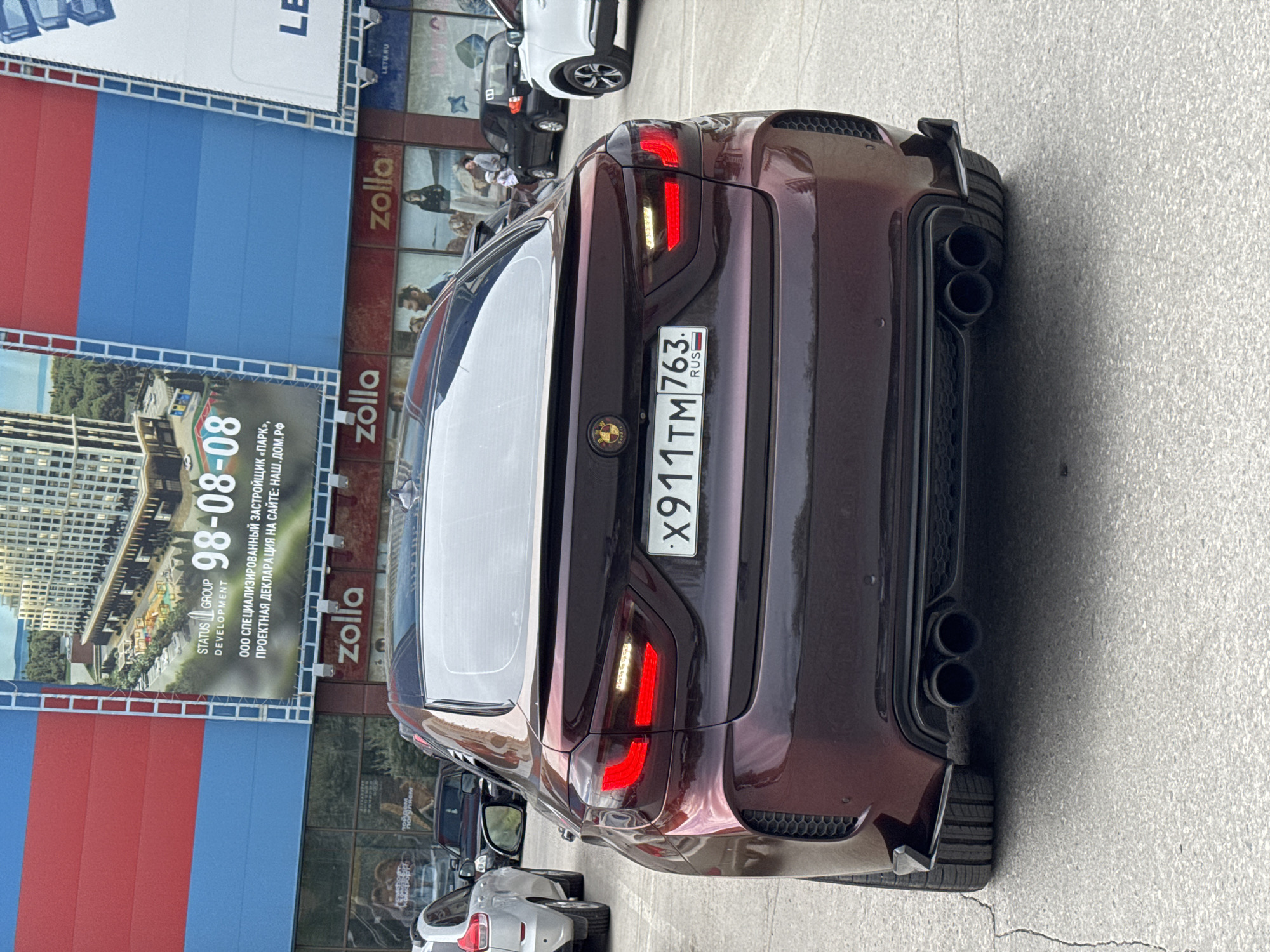 A rear view of a maroon car with a Russian license plate reading "X911TM 763 RUS" is shown. The car has quad exhaust pipes and modified taillights. The background reveals the facade of a building with the "zolla" logo and an advertisement for "STATUS GROUP DEVELOPMENT". The building appears to be located in a city of Russia. Other cars are parked in the background.