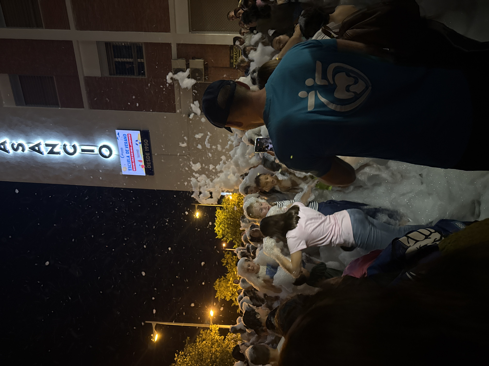 Here is a detailed description of the image:
**General Scene:**
The image depicts an outdoor event at night. A crowd of people are gathered, covered in foam, suggesting a foam party or similar activity.
**Location:**
A building is visible in the background with a sign that reads "ASANCIO" and "ESCUELA DE VERANO" (Summer School). The sign indicates the location may be a school. The text "DESDE 1950" suggests the establishment has been around since 1950. The address of the location is Unknown, Unknown.
**People:**
- There are numerous people of different ages, mostly youth, who are covered in foam.
- Some individuals are wearing casual clothing. One person is wearing a blue shirt with a white logo on the back.
- One person is holding a smartphone and appears to be recording or taking a photo of the scene.
**Objects:**
- Streetlights are present, illuminating the area.
- The building is of brick construction, as can be seen in the background.
- The foam appears to be artificially generated.
**Actions:**
- People are interacting, playing, and enjoying the foam.
- One person is actively recording the event with a smartphone.
**Environment:**
- It is nighttime.
- The environment is filled with artificial foam, suggesting a planned event.
- Streetlights are present, illuminating the event.