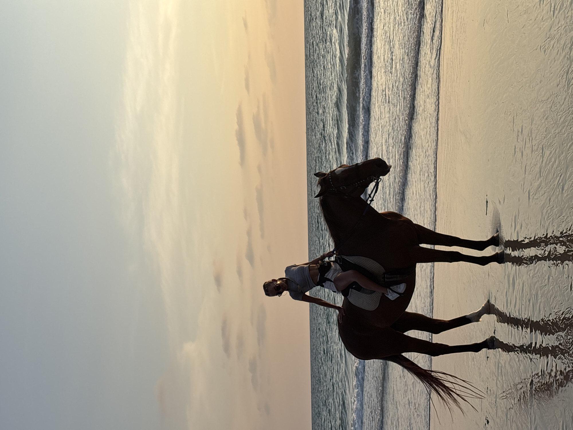 A woman rides a horse on a beach at sunset. The horse is brown, and the woman is wearing a light-colored top and shorts. The sky is a mix of orange, yellow, and blue, and the water is calm. The photo was taken on Bang Tao Beach in Choeng Thale, Thailand. The horse is partially submerged in the water, reflecting on the wet sand. The woman is leaning slightly back in the saddle.