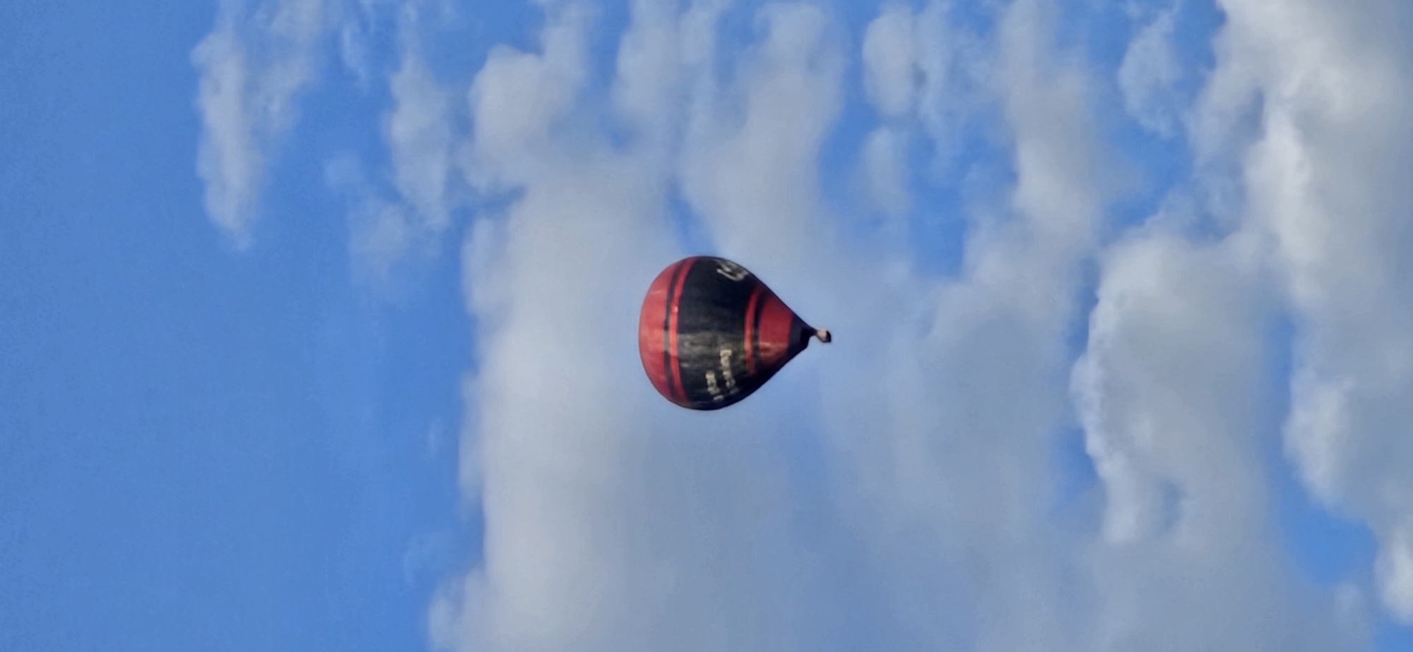 A single hot air balloon, predominantly red and black with white lettering that reads "The Amazing", ascends into a bright blue sky dotted with fluffy white clouds. The balloon is positioned centrally in the frame, appearing to be in mid-flight. The image captures a clear, sunny day. The location is specified as Hévíz, Hungary.