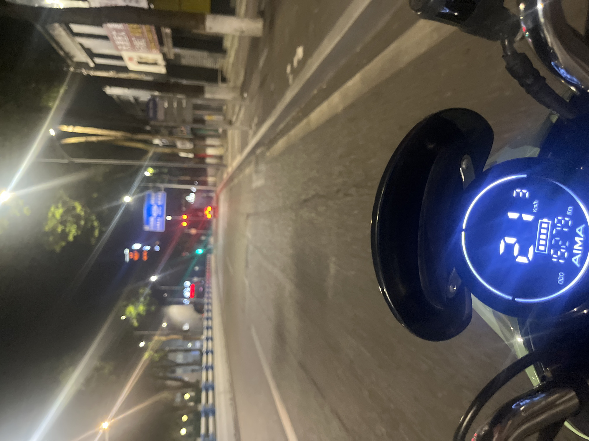 The image captures a nighttime urban street scene from the perspective of an electric scooter rider in Jiulongpo District, China. In the immediate foreground, the digital dashboard of an AIMA-brand scooter is visible, displaying a speed of 21 Km/h, an odometer reading of 1527.9 Km, and a nearly full battery indicator. Ahead, a multi-lane asphalt road stretches into the distance, illuminated by numerous streetlights on both sides. On the left, a median barrier separates the lanes, with more streetlights and trees beyond. On the right, commercial buildings with illuminated signs, some featuring Chinese characters, line the street. Traffic lights are visible further down the road, displaying both red and green signals. The taillight of a distant vehicle, likely a car, is visible in the center lane. The overall scene depicts a typical busy street environment at night. No individuals are explicitly visible.