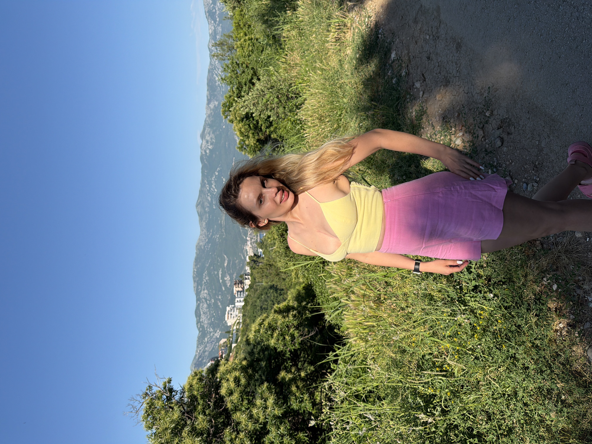 A woman is standing in a grassy area in Budva, Montenegro. She is wearing a yellow tank top and a pink skirt. In the background are mountains and the city of Budva. The sky is blue. There is a dirt path and some vegetation in the foreground. She is sticking her tongue out.