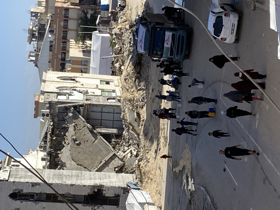The image depicts a street scene in Khan Yunis, Palestinian Territory, characterized by significant structural damage and humanitarian activity. In the background, a large, multi-story building with a light-colored facade, featuring arched windows on its intact sections, displays severe collapse in its central portion, leaving a substantial pile of rubble and exposed structural elements. Adjacent to this, other buildings appear damaged or partially ruined, and a large white tent is visible amidst debris.
In the foreground, a group of approximately 25-30 individuals, comprising men, women, and children, are present on an asphalt street with visible wet patches. Many carry or are waiting with plastic water containers, including jerrycans and buckets. A blue-green Volvo truck, bearing "Médecins Sans Frontières" (Doctors Without Borders) branding on its cab and a container in its bed, is parked on the right side of the street, with several people gathered around its rear, suggesting aid distribution. A white Nissan Micra-type vehicle is parked further down the street on the right. Utility lines crisscross the upper portion of the frame under clear daylight conditions.