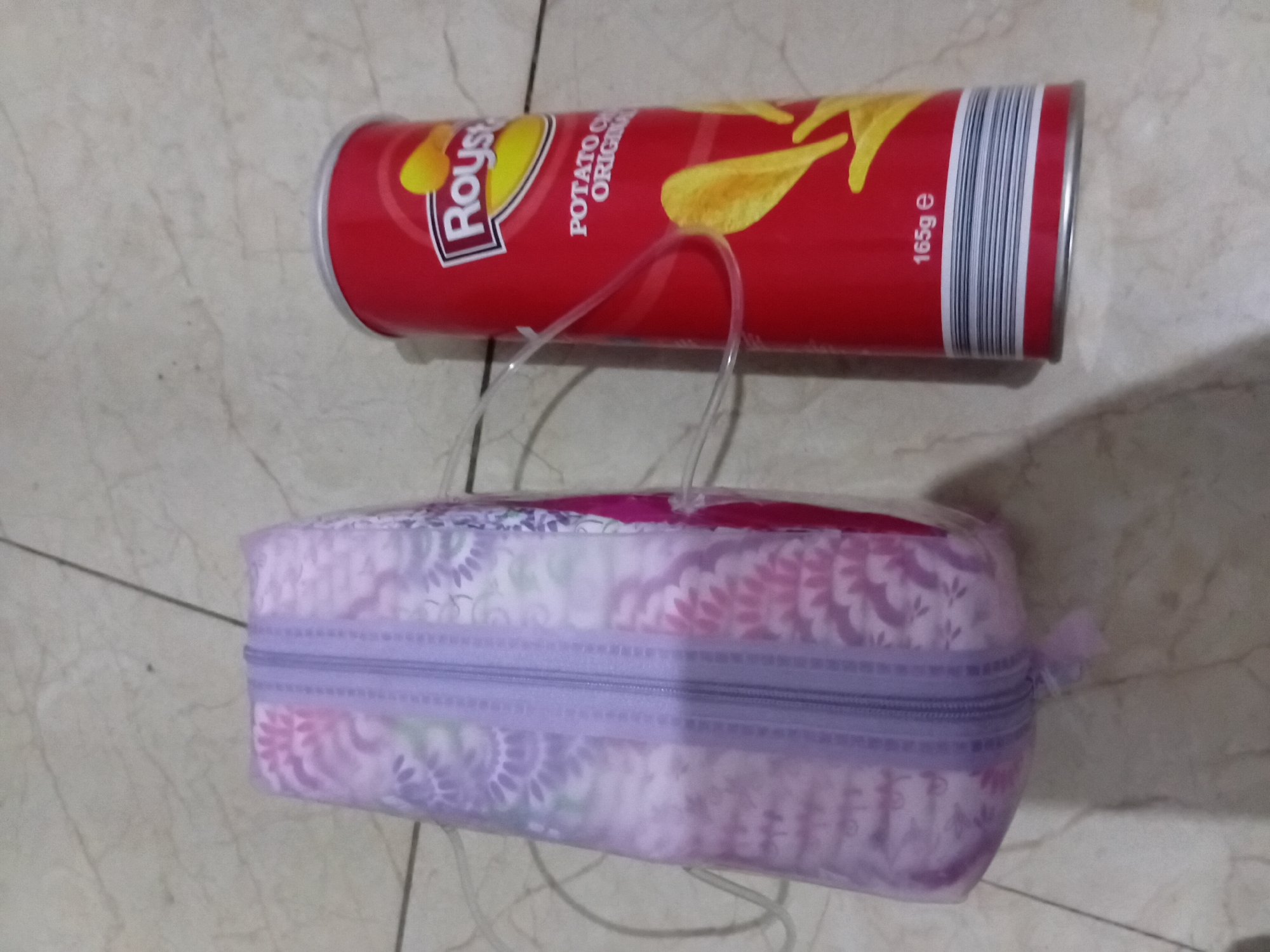 The image shows a can of Pringles-like chips labeled "Roys Original Potato Chips 165g e" lying on a patterned, light-colored floor, along with a rectangular zippered pouch with a floral pattern and a clear plastic handle. The location is Akwete, Nigeria.