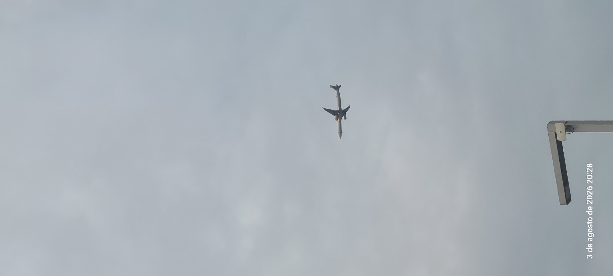 A commercial airplane is captured mid-flight against a cloudy sky. The plane is white with yellow accents on its engines and tail fin, and its landing gear is extended, suggesting it is either approaching or departing an airport. In the foreground, a portion of a street lamp or similar urban lighting fixture is visible. The timestamp "3 de agosto de 2026 20:28" is overlaid on the bottom left of the image. The scene appears to be an outdoor urban environment, possibly near an airport.