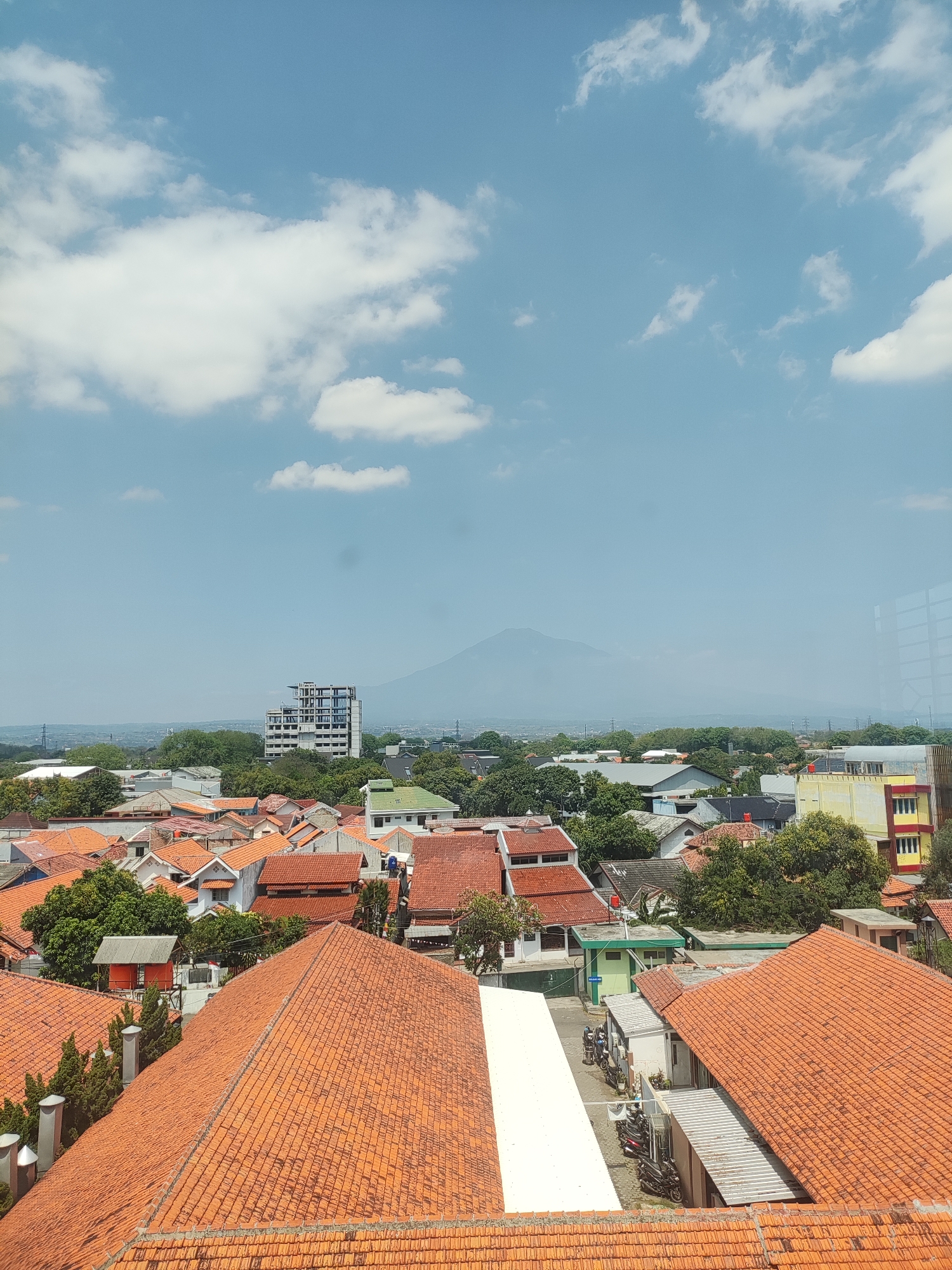 The image shows an aerial view of a residential area in Cirebon, Indonesia, with a mountain in the distance. The foreground is dominated by red-tiled roofs of houses, some with multiple stories. Trees are interspersed among the buildings, providing greenery. In the mid-ground, a modern multi-story building stands out, and further in the distance, a mountain is faintly visible through a hazy atmosphere. The sky is a clear blue with scattered white clouds. The scene depicts a typical urban landscape with a blend of residential structures, vegetation, and natural elements.