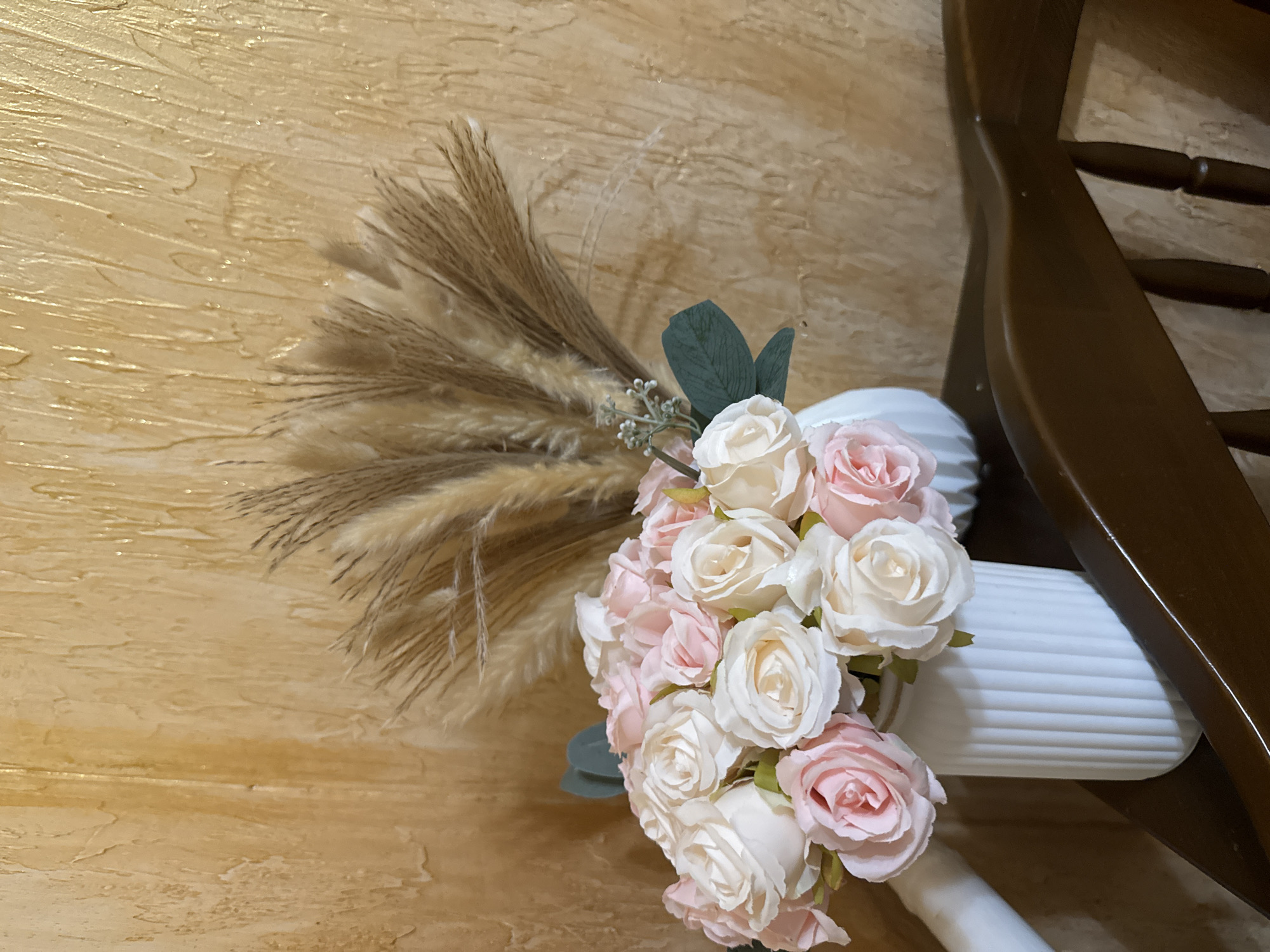 An arrangement of light pink and cream roses and dried Pampas grass is displayed in white vases against a textured, golden-brown wall. The flowers are artificial and arranged in a way that suggests a decorative piece. The Pampas grass is tall and feathery, contrasting with the softer texture of the roses. The wall has a mottled, somewhat distressed appearance, with streaks of lighter and darker tones. The foreground features the dark brown wood of a piece of furniture, possibly a chair or table. The lighting is soft, creating a warm atmosphere.