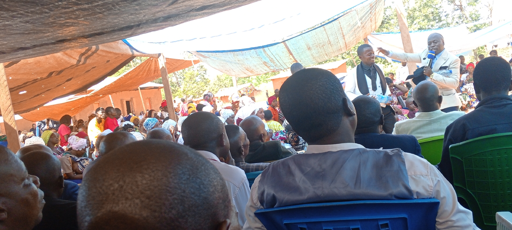 Under a makeshift canopy in Kalemie, Democratic Republic of the Congo, a crowd has gathered. Dominating the foreground are the backs of heads belonging to seated attendees. An adult man in a light-colored suit stands, holding a microphone and gesturing, presumably addressing the gathering. A second man, in a black suit, stands beside him. The assembled group is diverse, with individuals of various ages and attire. Structures, possibly dwellings, are visible in the background, suggesting a residential area. The scene is bathed in natural sunlight, with shadows cast by the canopy.