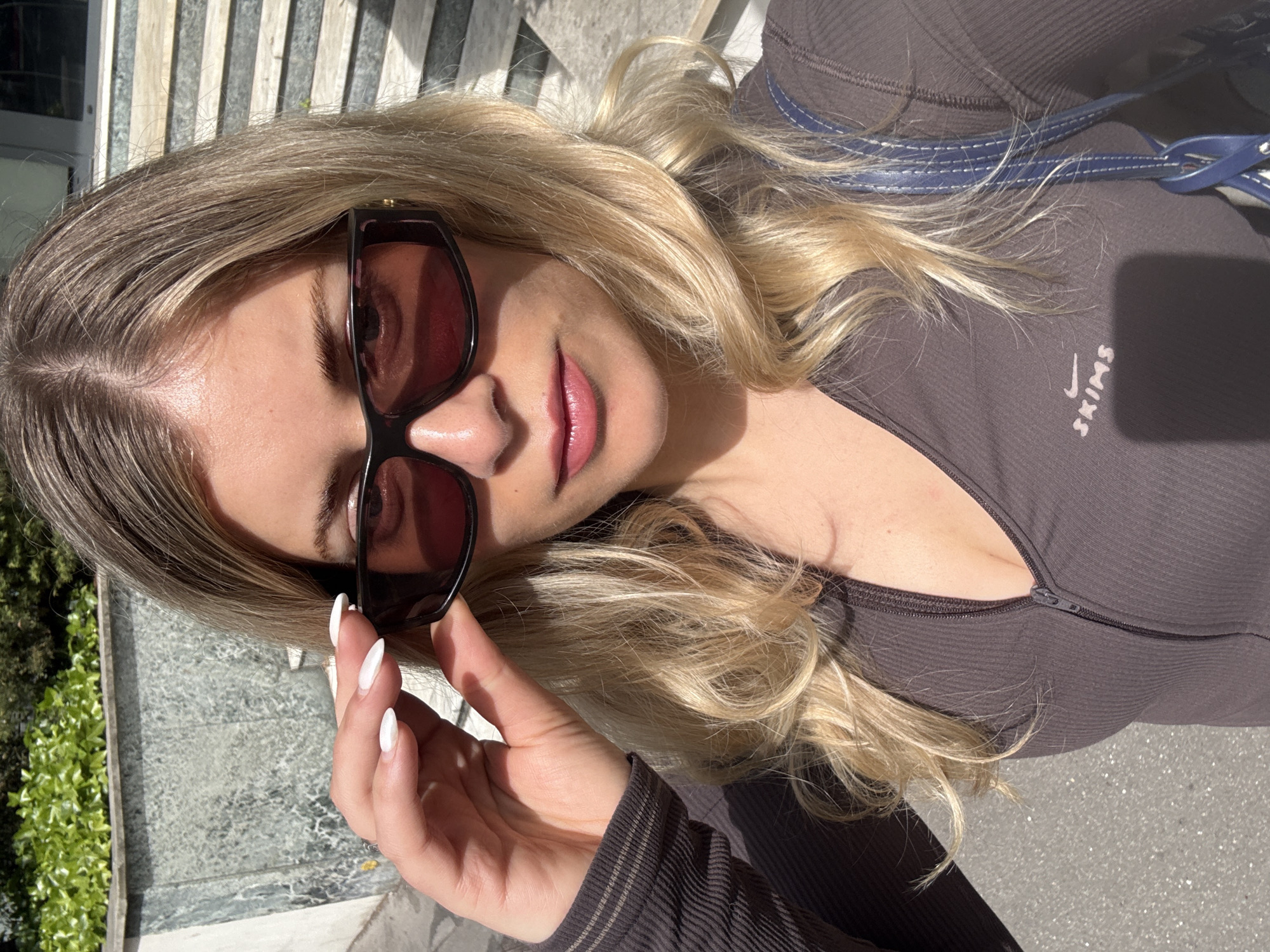 A woman with blonde hair and fair skin is captured in a close-up selfie, tilting her head slightly. She wears large, dark rectangular sunglasses, one of which she is holding with her right hand, revealing long white manicured nails. Her lips are adorned with a glossy pinkish-red lipstick. She is dressed in a fitted, dark brown ribbed top with a zipper, and on her chest, a light-colored "SKIMS" logo with a small swoosh is visible. A blue strap with white stitching, possibly from a bag, rests on her shoulder.
The image is taken outdoors in bright, sunny conditions, casting strong shadows that highlight the textures of her hair and clothing. The background, largely blurred, shows glimpses of green foliage on the left and a light-colored, possibly paved or stone, surface. Behind her head, a light-colored slatted structure, possibly a bench or railing, can be seen. The overall setting suggests a pleasant daytime outdoor environment in Paris, France. The woman appears relaxed, looking towards the camera through her sunglasses.