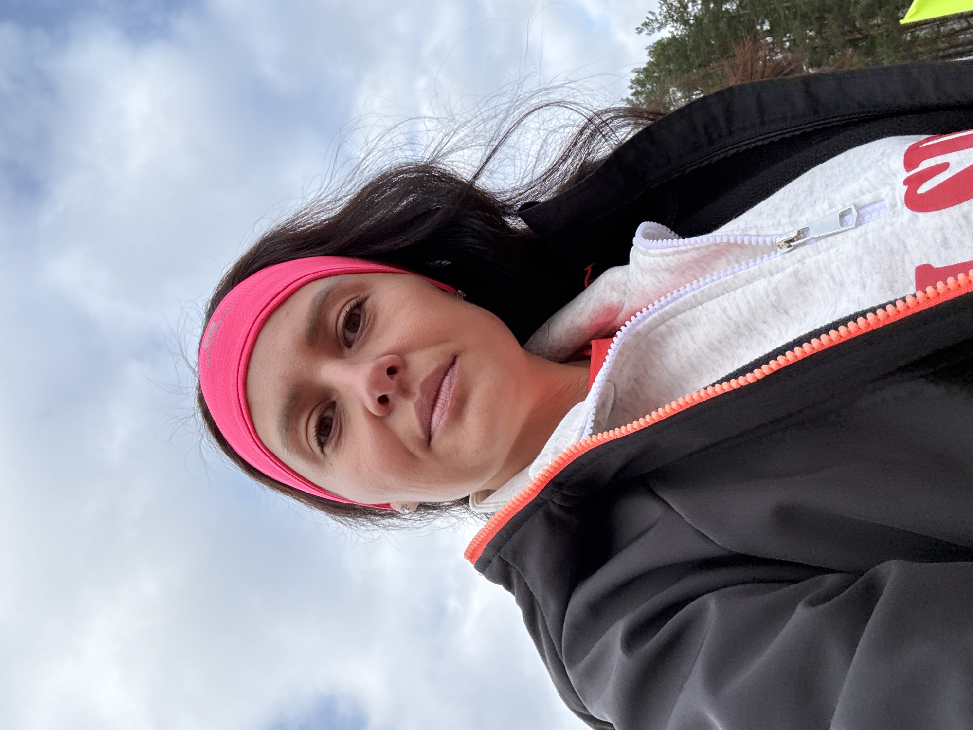 A woman is taking a selfie outdoors under a cloudy sky. She is wearing a pink headband, a white zip-up hoodie with red writing on it, and a black jacket with a bright orange trim along the zipper. The location and city of Chaikovsky, Russia, cannot be determined from the image.