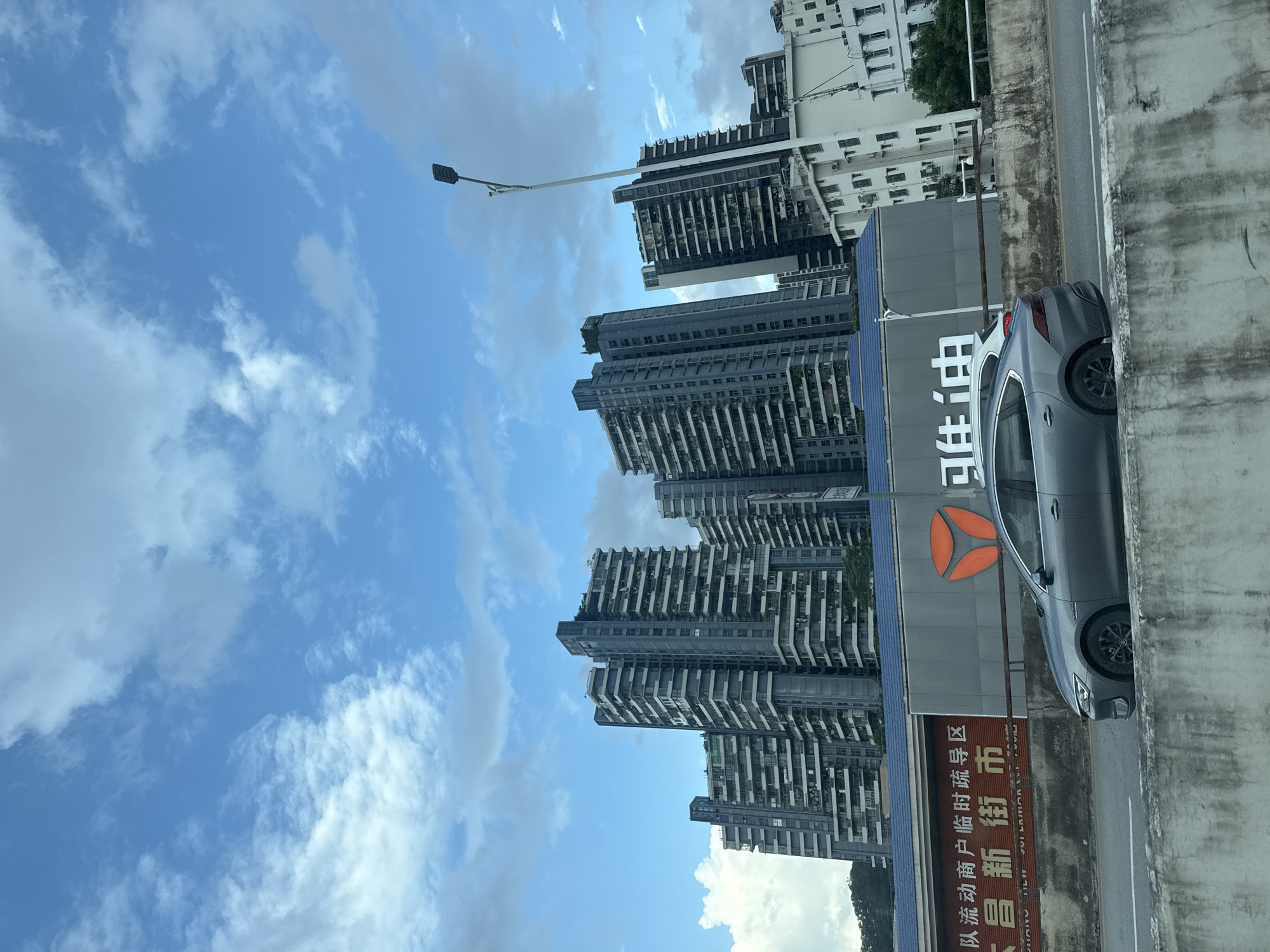 This image captures a scene in Huizhou, China, featuring a series of modern, high-rise apartment buildings against a backdrop of a blue sky with scattered white clouds. In the foreground, a grey sedan is driving on a road bordered by a concrete barrier. Below the apartment buildings, there is a long, grey building with a blue roofline and signage. One prominent sign displays Chinese characters and an orange logo. To the right, another white building with multiple windows is visible, and a street light stands tall. The overall impression is one of urban development in a well-populated area.