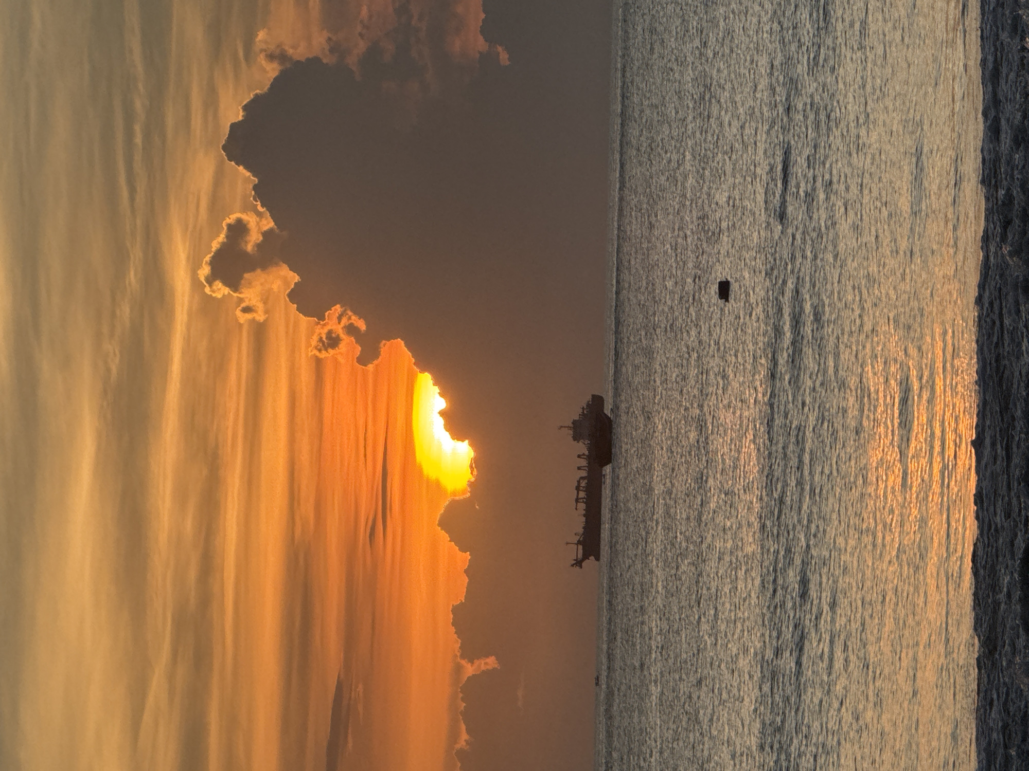 The image captures a sunset scene in Kuta, Indonesia, with the sun partially obscured by clouds. The sky is awash in orange and yellow hues, reflecting on the surface of the ocean. A cargo ship is visible on the horizon. A small dark object floats in the sea between the ship and the shore. The shoreline is visible in the bottom right corner.