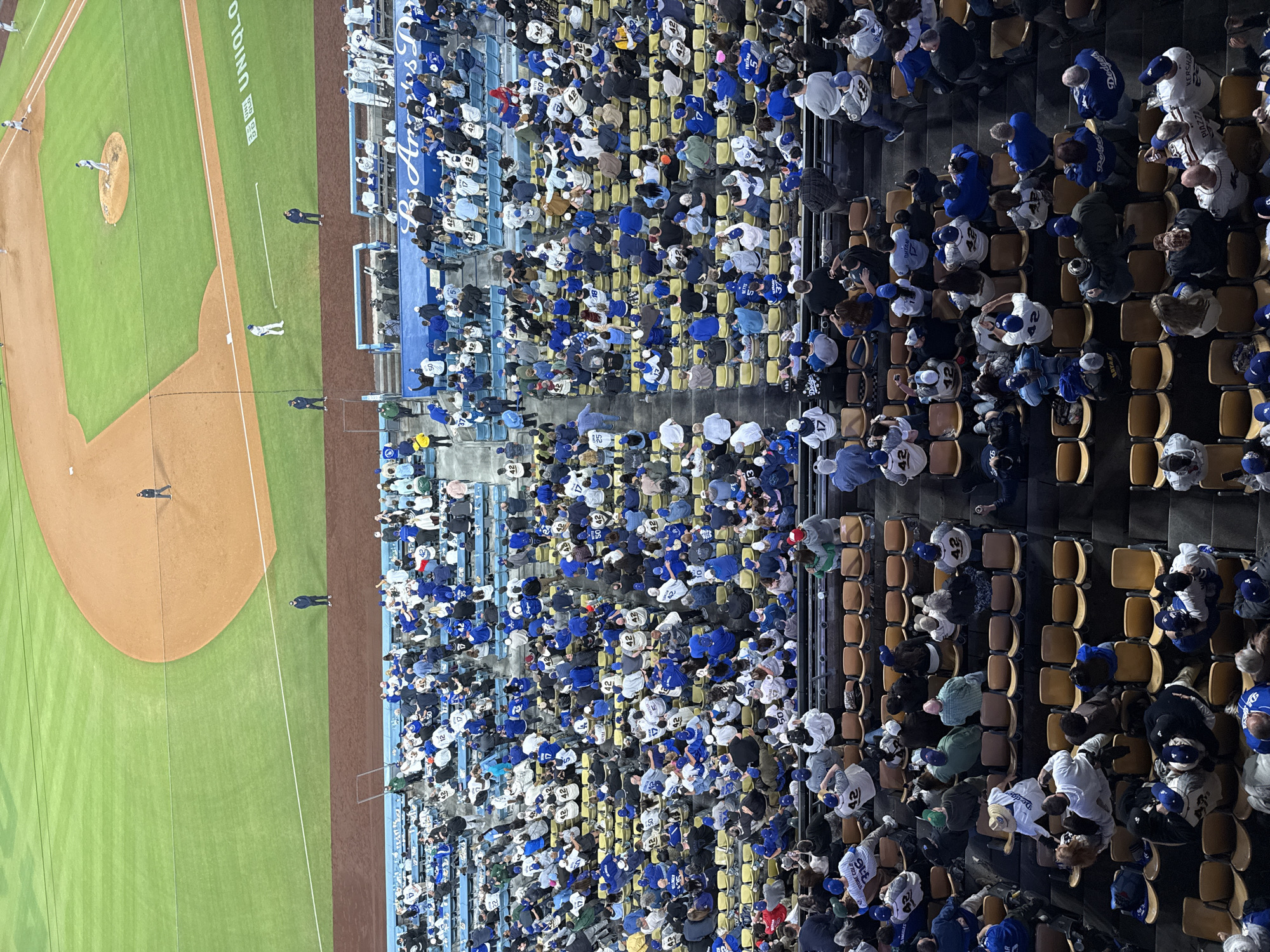 The image depicts an aerial view of a baseball game taking place at Dodger Stadium, located in Los Angeles, United States of America. The game is in progress, with players visible on the field. The stands are filled with a large crowd of spectators, many of whom are wearing Los Angeles Dodgers apparel, primarily in white and blue colors. The crowd is seated in tan-colored seats. Various advertisements are displayed, including one that reads "UNIQLO." The environment suggests a night game setting due to the lighting conditions.