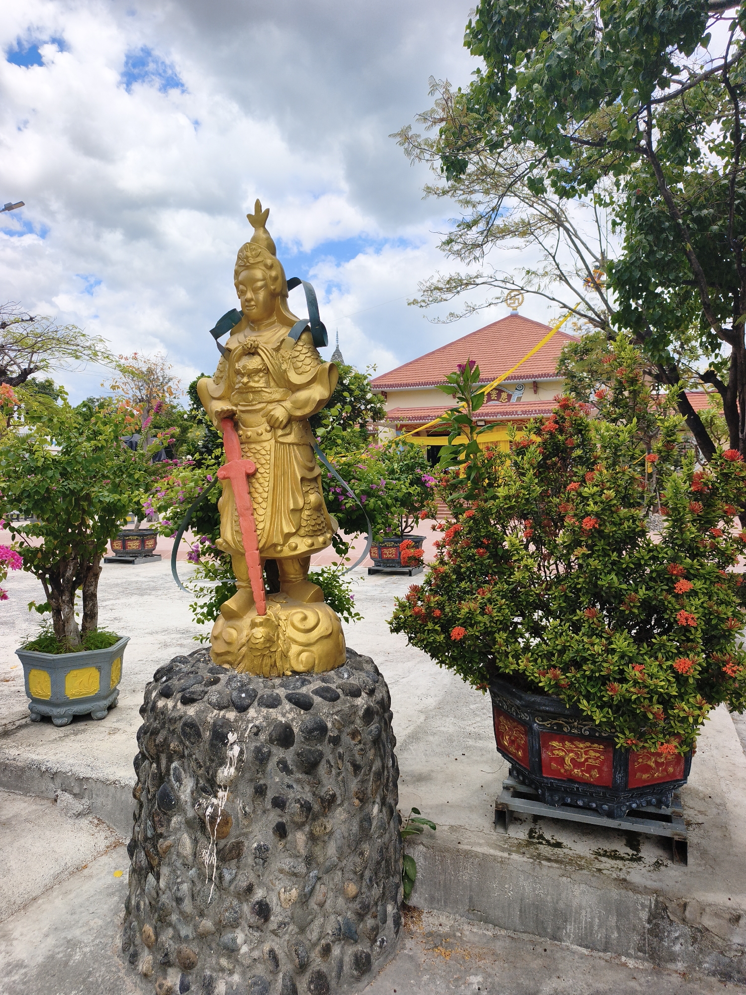 A golden statue of a warrior stands on a rough, stone pedestal in front of a building with a red tiled roof. The statue holds a red sword and is adorned with armor and a headdress. The scene is outdoors, with lush green trees and flowering bushes surrounding the statue and building. The sky is cloudy with patches of blue. The building appears to be a temple or a similar religious structure. The statue is likely a representation of a deity or a historical figure, common in Buddhist or Taoist traditions. The overall atmosphere suggests a serene and spiritual setting, possibly within a temple complex in Cam Ranh, Vietnam.