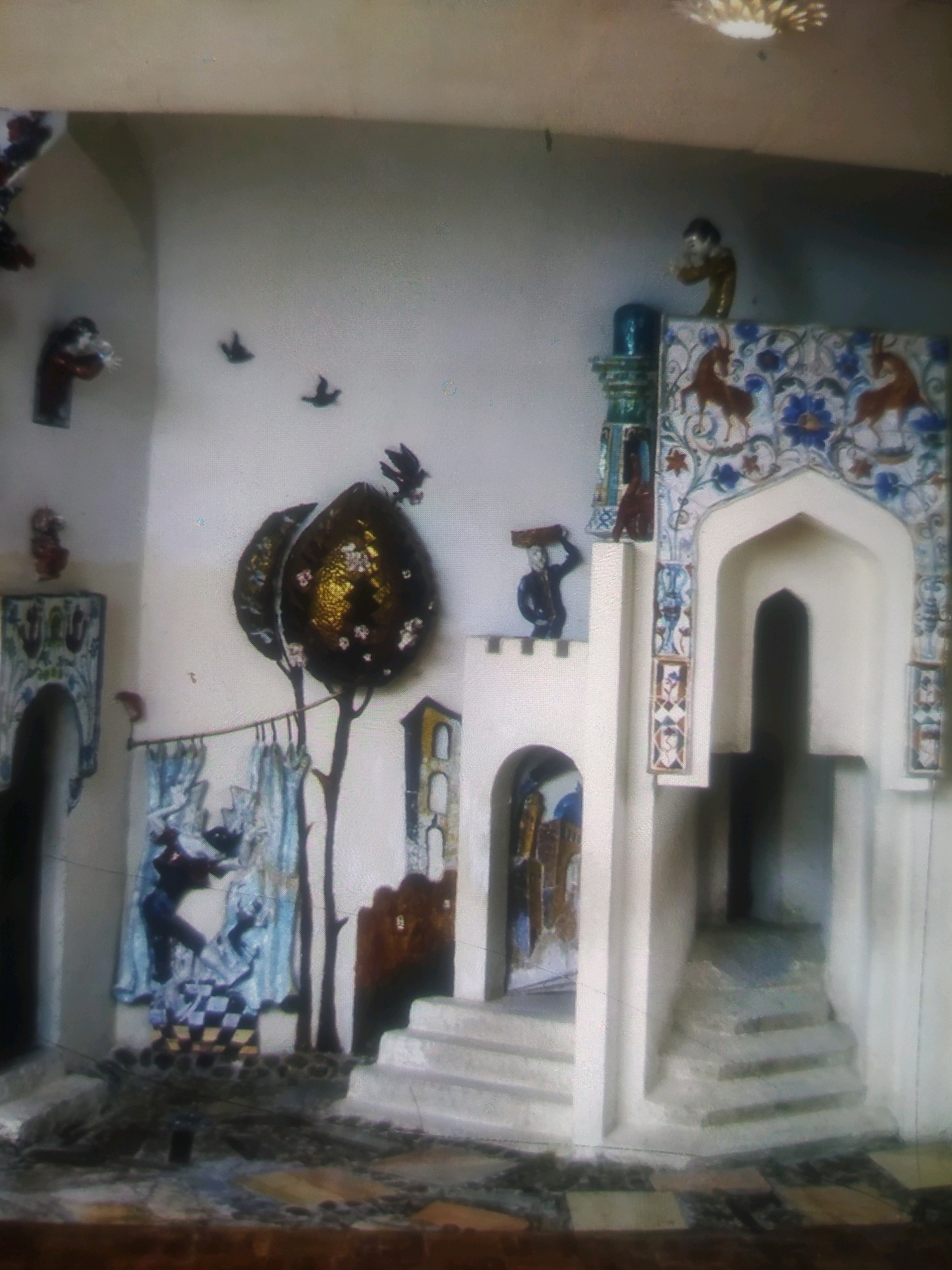 The image depicts an indoor scene, possibly a museum or artistic installation, featuring a collection of whimsical, stylized decorative elements on a pale gray wall. To the left, an archway with decorative blue and green tilework frames an opening. Above and to the left of this archway, several small, dark bird figures appear to be flying or perched. A large, dark, tear-drop shaped object with a textured, gold-leafed surface, resembling stylized fruit or pods on a thin, dark tree, dominates the mid-left of the frame. To the right of this, a small, yellow, tower-like structure with arched openings is visible.
Further to the right, a white, fortress-like structure rises, topped with a small figure of a person carrying a red object on their head. Adjacent to this is an elaborate, arched alcove, decorated with colorful mosaic tiles featuring red animals that appear to be goats or deer, and blue floral motifs. Steps lead up to this alcove. A small, dark figure is visible at the top of the alcove. Below the tiled alcove and to the left, a decorative panel depicts figures in blue and white against a checkered pattern. The floor is paved with stones of various colors and textures. A subtle, circular light fixture is visible on the ceiling in the upper right quadrant of the image. There is no indication of the specific location within the city of Birlik, Uzbekistan.