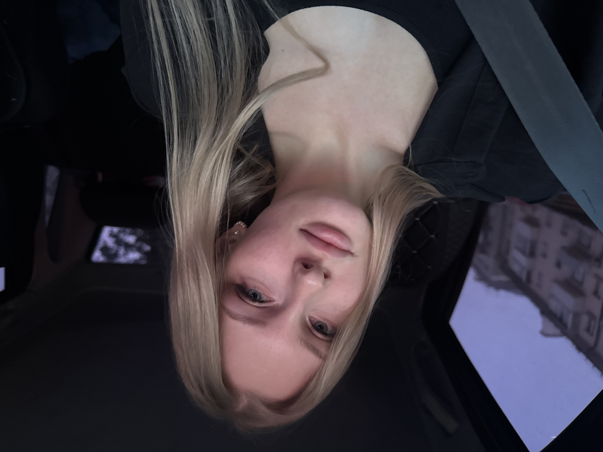 A light-skinned, young woman with blonde hair is in a car. She is wearing a black top and a seatbelt is visible. The photo is taken from a slightly low angle, facing upwards. Outside the car window, a building is visible, suggesting an urban environment. The building is blurry, making it difficult to identify specific details. Given the information provided (city of Perm, Russia), it is likely the image was taken there.