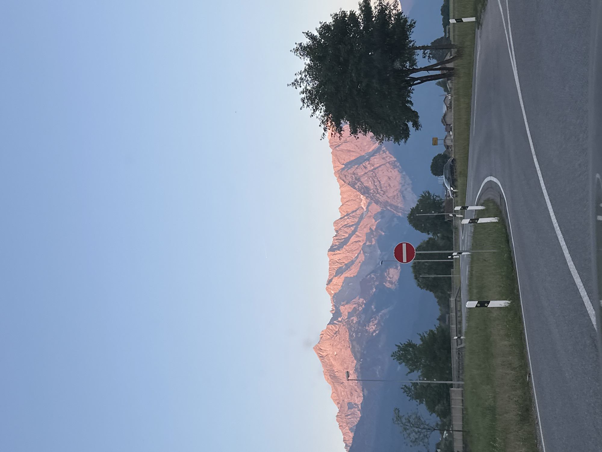 A scenic view of the Alps, bathed in the pink glow of sunset, dominates the background of this image taken near Garmisch-Partenkirchen, Germany. The mountains, with their jagged peaks, stretch across the horizon. In the foreground, a paved road curves to the right, bordered by grassy verges and white-and-black bollards. A single, large tree stands on the right side of the road. A red and white "no entry" sign is visible on a pole. The sky is a clear, pale blue, transitioning to a warmer hue near the mountains. A few scattered trees and buildings can be seen in the middle ground, along with a dark-colored car parked on the side of the road. The lighting suggests it is either early morning or late evening.