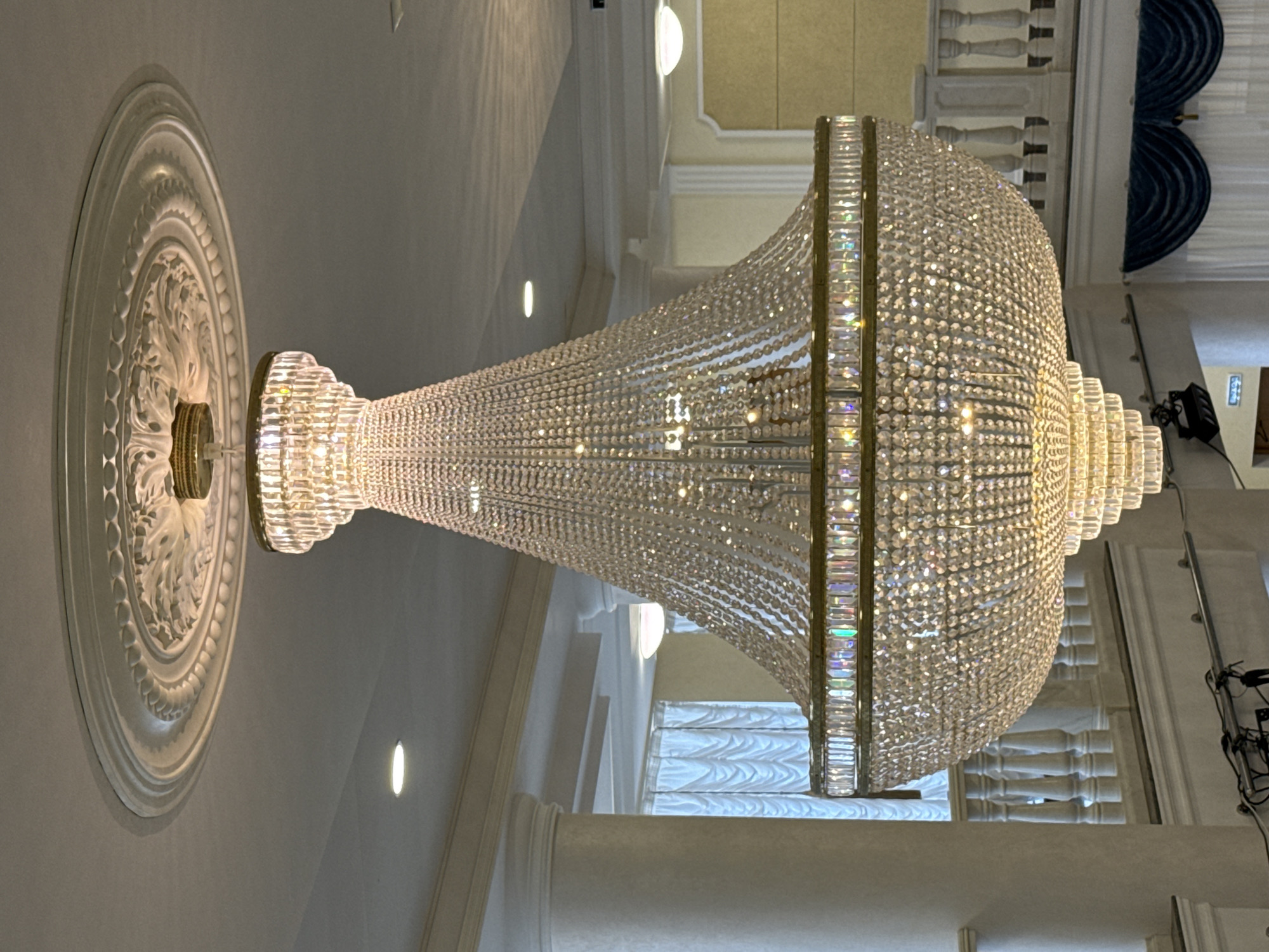 A chandelier is suspended from a decorative ceiling fixture. The chandelier is large and made of crystals. The surrounding architecture is white and ornate. The location is indeterminate.