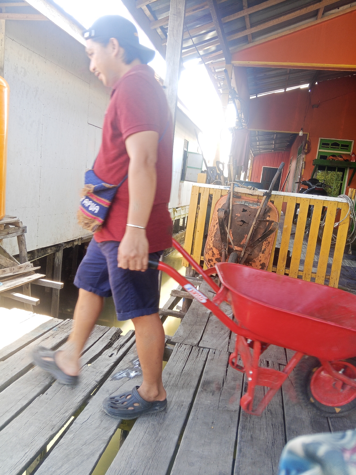 In Tarjun, Indonesia, a person is pushing a red wheelbarrow across a wooden pier. They are wearing a maroon polo shirt, navy blue shorts, dark grey perforated clogs, and a black baseball cap. A small, colorful woven bag with fringe is slung across their body. The pier is constructed from weathered grey wooden planks, and the person is walking towards the right side of the frame. Sunlight streams from the left, creating bright highlights. In the background, there are wooden stilt houses and a structure with orange walls and a yellow fence. Tools, including another wheelbarrow, are visible near the fence. The scene suggests an outdoor, possibly residential or work area, with a focus on manual labor or transport.