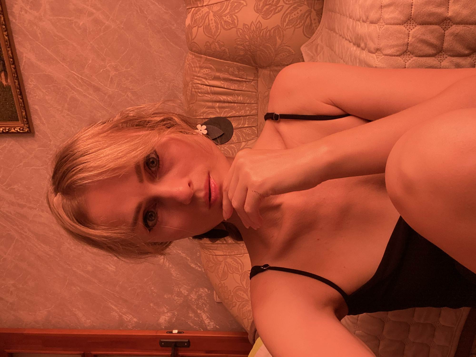 A young woman with blonde hair and dark eyeliner is pictured in an indoor setting. She is wearing a black camisole and large, dark, floral-shaped earrings. Her right hand is resting on her chin, and she is looking directly at the camera. The background appears to be a wall with a textured pattern, and behind her is a sofa with a patterned fabric. A framed picture is visible on the wall above the sofa. The lighting in the image casts a strong orange or red hue over the scene, possibly from an artificial light source. The overall impression is a close-up, posed portrait. There is no information in the image to suggest a specific location in Lida, Belarus.