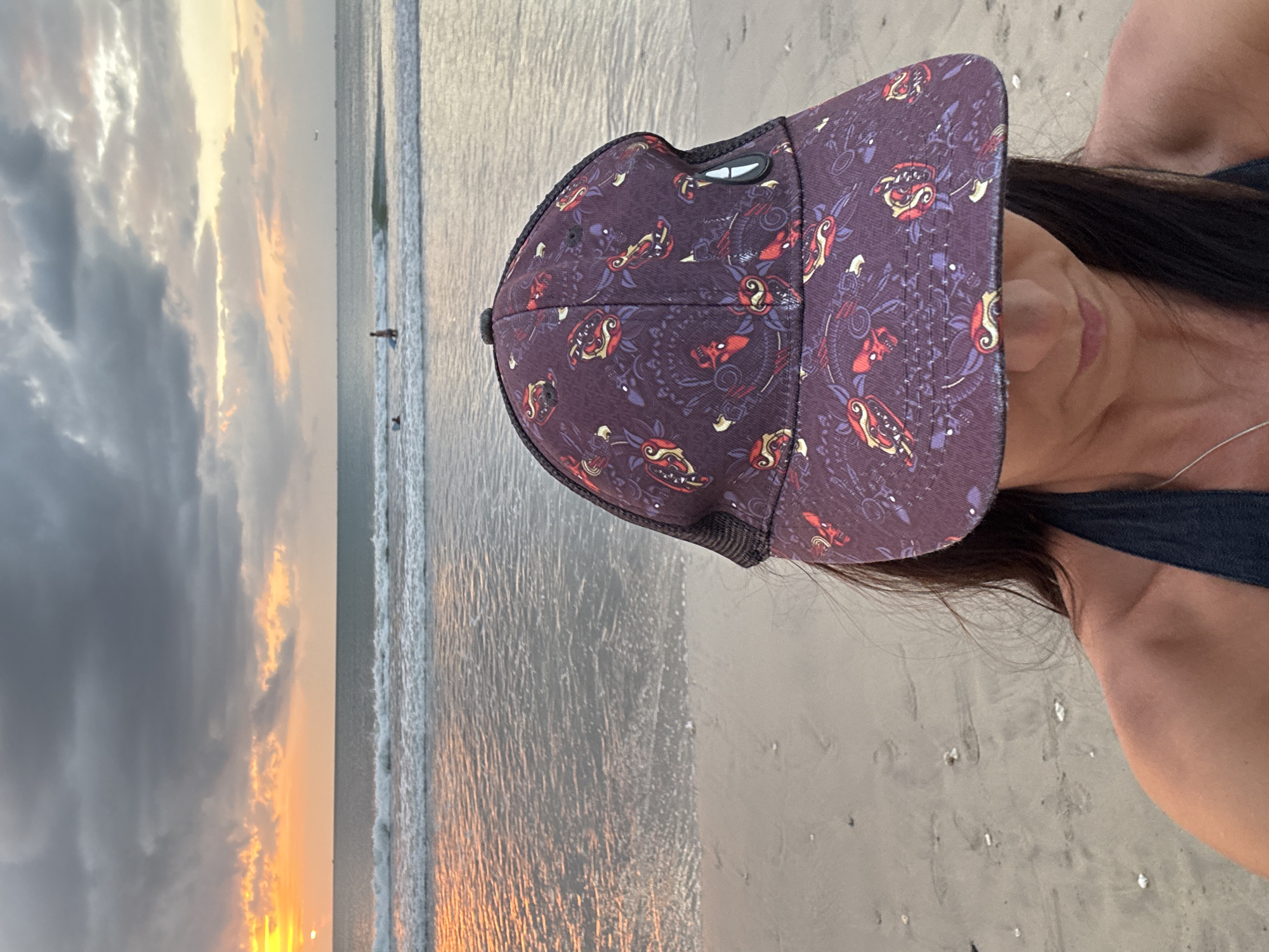 An over-the-shoulder selfie captures a woman wearing a patterned cap and a dark bikini top against a beach and ocean backdrop at sunset. The sky is dramatic with dark, heavy clouds interspersed with areas of orange and yellow light, indicating the sun is low on the horizon. Waves gently roll onto a sandy beach. In the middle distance, several figures can be seen in the water, some appearing to be surfing or wading. A small airplane is visible in the far distance, in the upper right quadrant of the image. The location is likely Kuta Selatan, Indonesia, known for its sunset beaches.