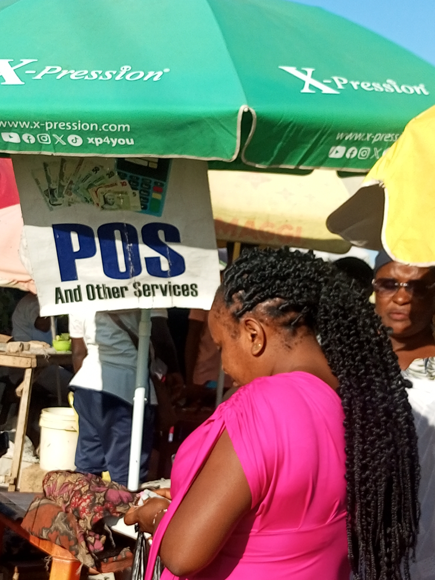 This image captures a bustling street scene in Tempure, Nigeria, likely near a market or commercial hub, given the presence of a vendor and various goods. Dominating the upper portion of the frame is a large, green umbrella, a common sight for shade in outdoor markets. The umbrella is branded with "X-Pression" in white, stylized lettering and also displays the website "www.x-pression.com" along with social media icons (Facebook, Instagram, Twitter, TikTok) and the handle "xp4you". Below the umbrella, a white sign proclaims "POS And Other Services" in large blue letters, with smaller text below it. Behind the sign, a collection of Nigerian Naira banknotes is visible, along with a calculator, suggesting a financial service point.
In the foreground, a woman with dark, braided hair pulled back into a long ponytail is the primary subject. She is wearing a vibrant, fuchsia pink top with a ruched detail on the shoulder. Her attention appears to be directed downwards, possibly at a transaction or personal device she is holding. To her right, partially obscured by her hair, is another person wearing glasses and a patterned top. Their gaze is also directed forward, suggesting they might be a customer or another vendor.
The environment appears somewhat cluttered, typical of a street market. To the left, a wooden structure and a white bucket are visible. The lighting suggests it is daytime, with sunlight casting shadows. The overall impression is one of commercial activity and daily life unfolding in an urban Nigerian setting.