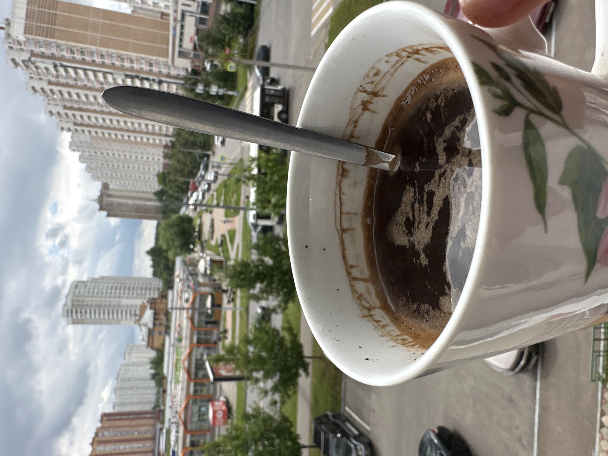 In the foreground, a white ceramic mug with a floral pattern is held by a person's hand. The mug contains dark coffee with a spoon submerged in it. In the background, a city street is visible with multiple cars, buses, and pedestrians. Tall apartment buildings and a strip of shops line the street. The sky is overcast. The scene appears to be set in the city of Podolsk, Russia, likely in a residential area with commercial establishments.