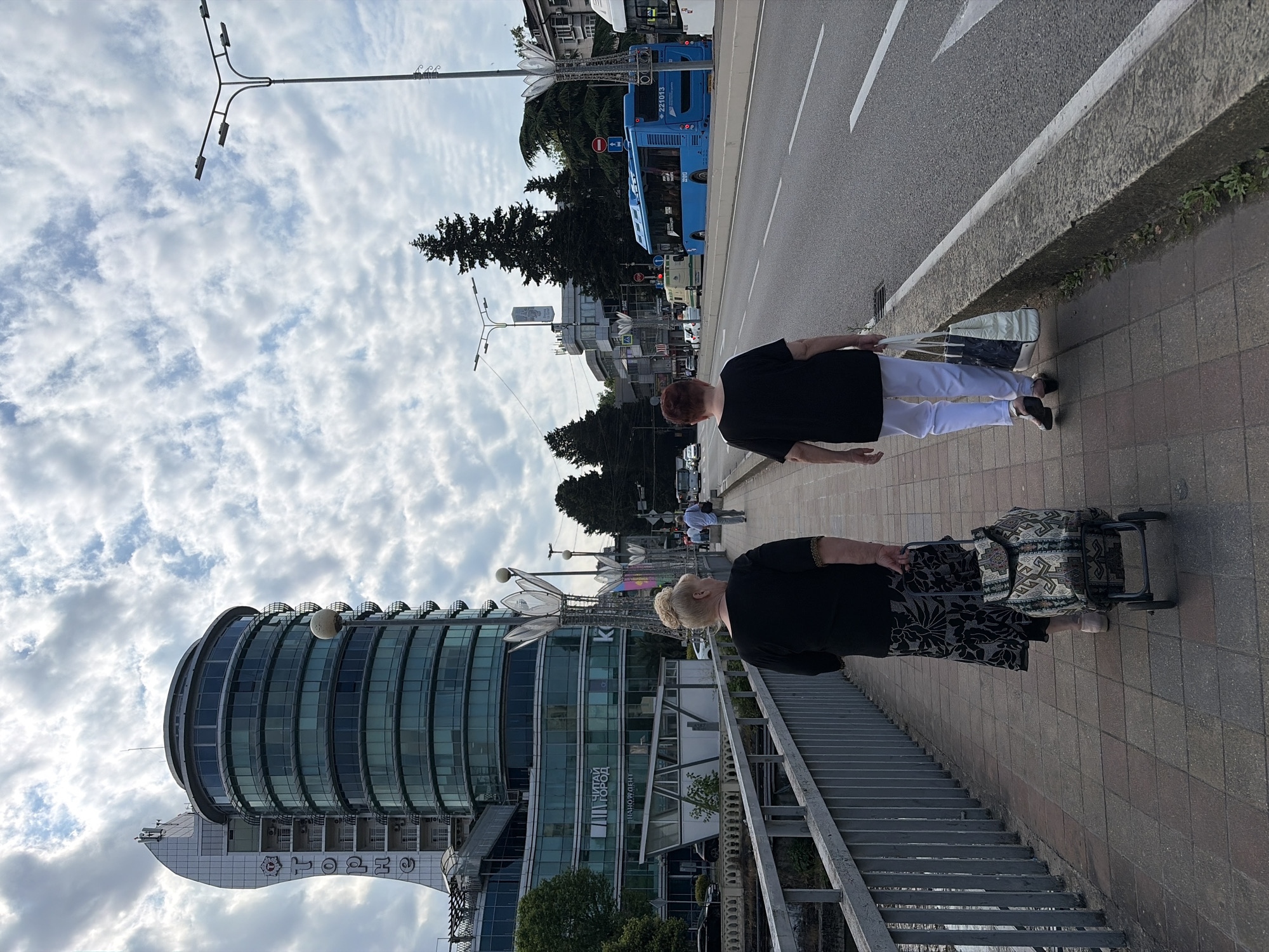 Two women are walking on a sidewalk next to a road in Sochi, Russia. The woman on the left is wearing a black shirt and a patterned skirt, and she is pulling a wheeled shopping cart. The woman on the right is wearing a black shirt and white pants, and she is carrying a bag. In the background, there is a modern, circular glass building, trees, streetlights, and a blue bus. The sky is cloudy.