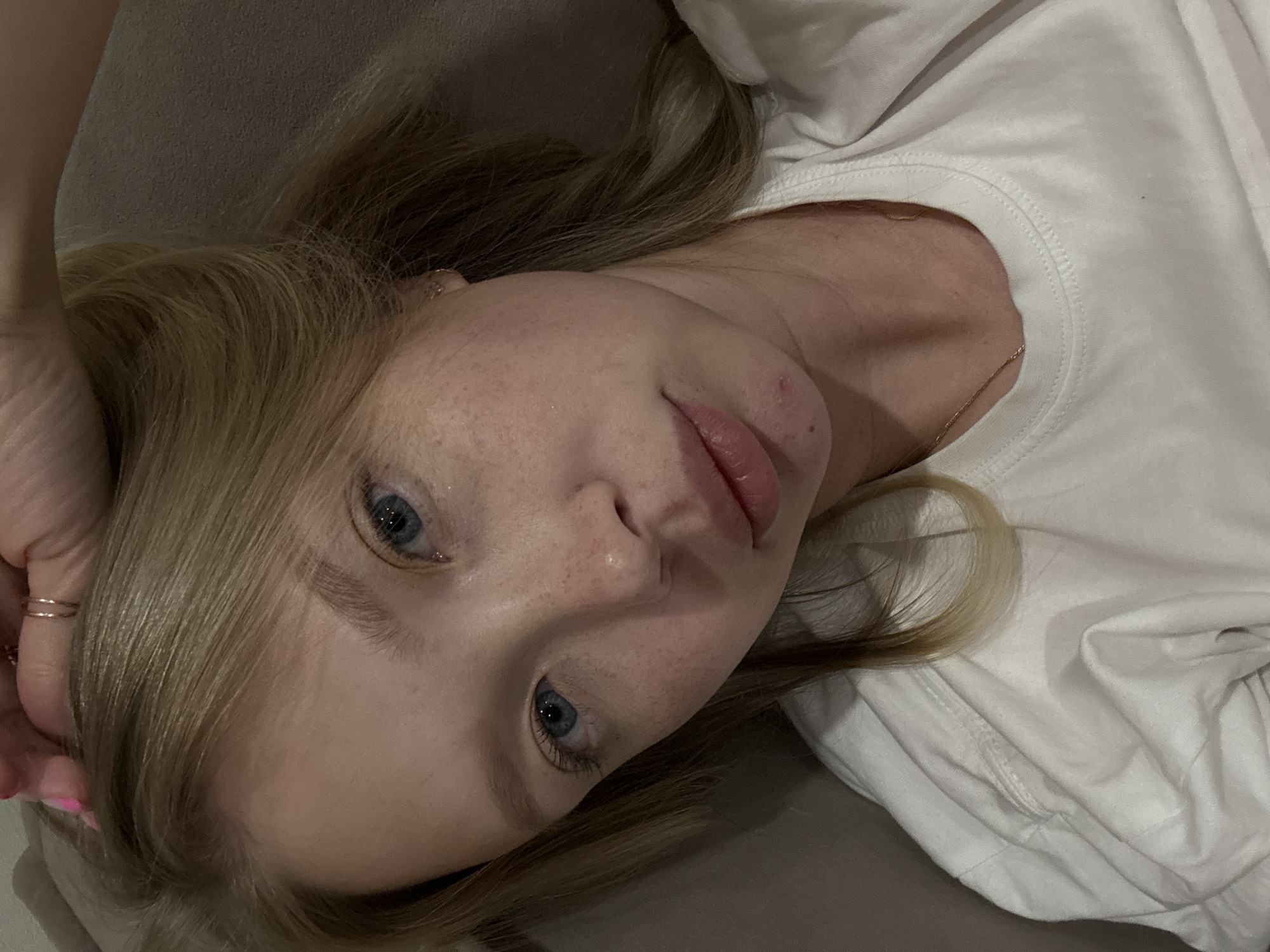 A young woman with blonde hair and blue eyes is lying down, holding her hand to her head. She is wearing a white t-shirt and a gold necklace. There are some freckles on her face and a pimple on her chin. The background is a blurred beige surface, possibly a couch. The image appears to be a selfie taken indoors. There is no identifiable information or context that places the scene in Chaikovsky, Russia.