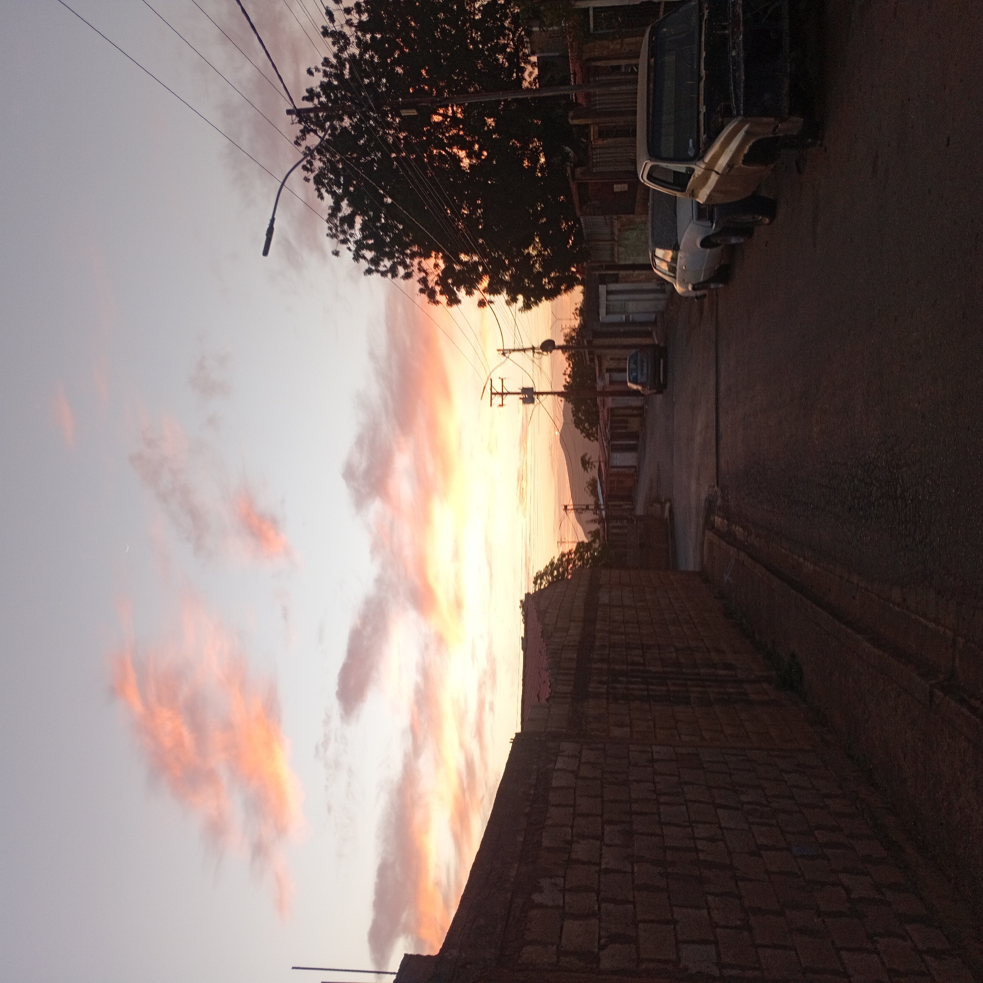The image shows a street scene at sunset in an unknown city. A brick wall runs along the lower right of the frame, and a street with parked cars is visible on the right side. Overhead power lines crisscross the sky, and a tree with dark foliage is present. The sky is filled with clouds colored in hues of orange and yellow from the setting sun. A faint crescent moon is visible in the upper left corner.