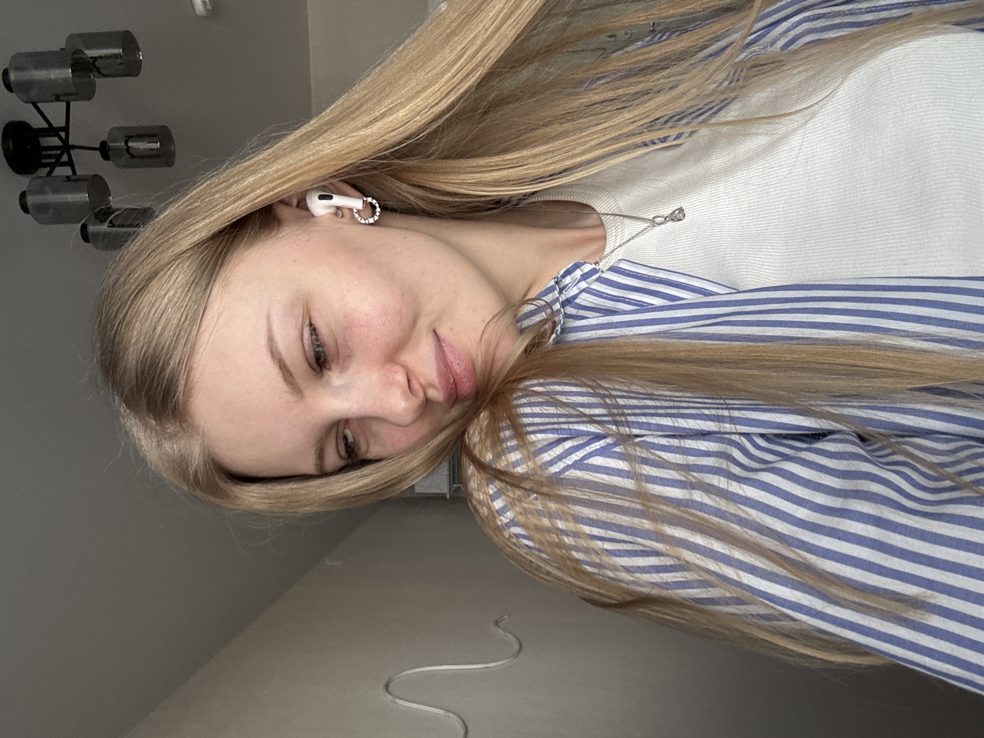 A blonde woman is indoors, wearing a white sweater under a blue and white striped shirt. She has an earbud in her ear and a silver necklace. The background consists of a gray wall and ceiling fixture. The photograph was taken indoors in Perm, Russia.