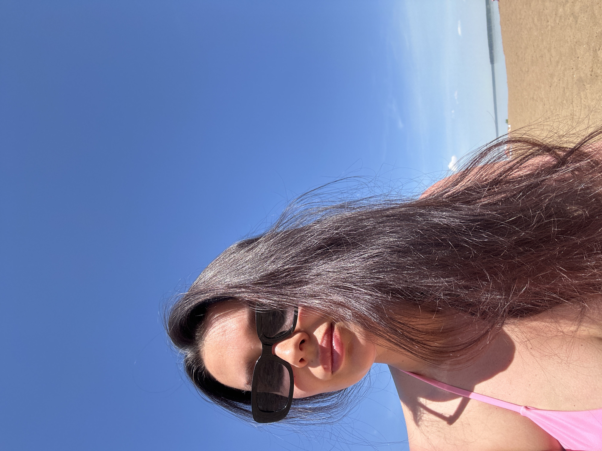 A medium shot depicts a woman on a beach in Chaikovsky, Russia. She has long, dark hair and wears dark sunglasses. She is wearing a pink bathing suit. The background shows a clear blue sky, and a strip of sand and water is visible in the distance.