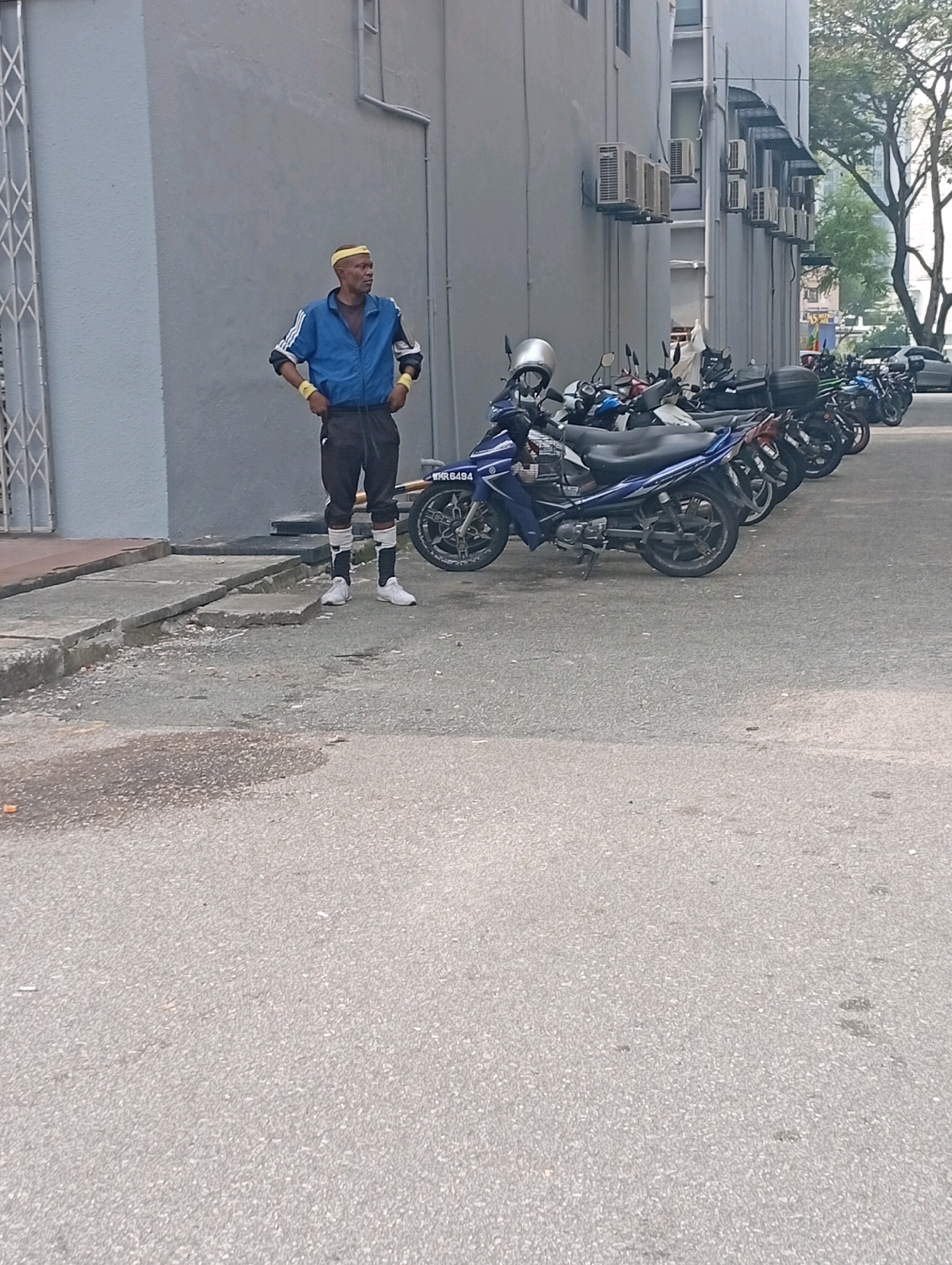A full-shot, eye-level, vertical, outdoor, natural light, daytime photograph captures a man standing on a cracked concrete pathway next to a row of parked motorcycles. The man, who appears to be of African descent, is wearing a blue athletic jacket with white stripes on the sleeves, black athletic pants with white stripes, white sneakers, and a yellow headband. His hands are on his hips, and he is looking to his right. The motorcycles are parked in a line on the asphalt, with a blue motorcycle prominently in the foreground. Behind the motorcycles is a building with grey walls and multiple air conditioning units mounted on the exterior. A tree with lush green foliage is visible in the background, suggesting a tropical or subtropical environment, consistent with the city of Selayang Municipal Council, Malaysia. The scene appears to be a public space, likely a parking area outside a commercial or residential building.