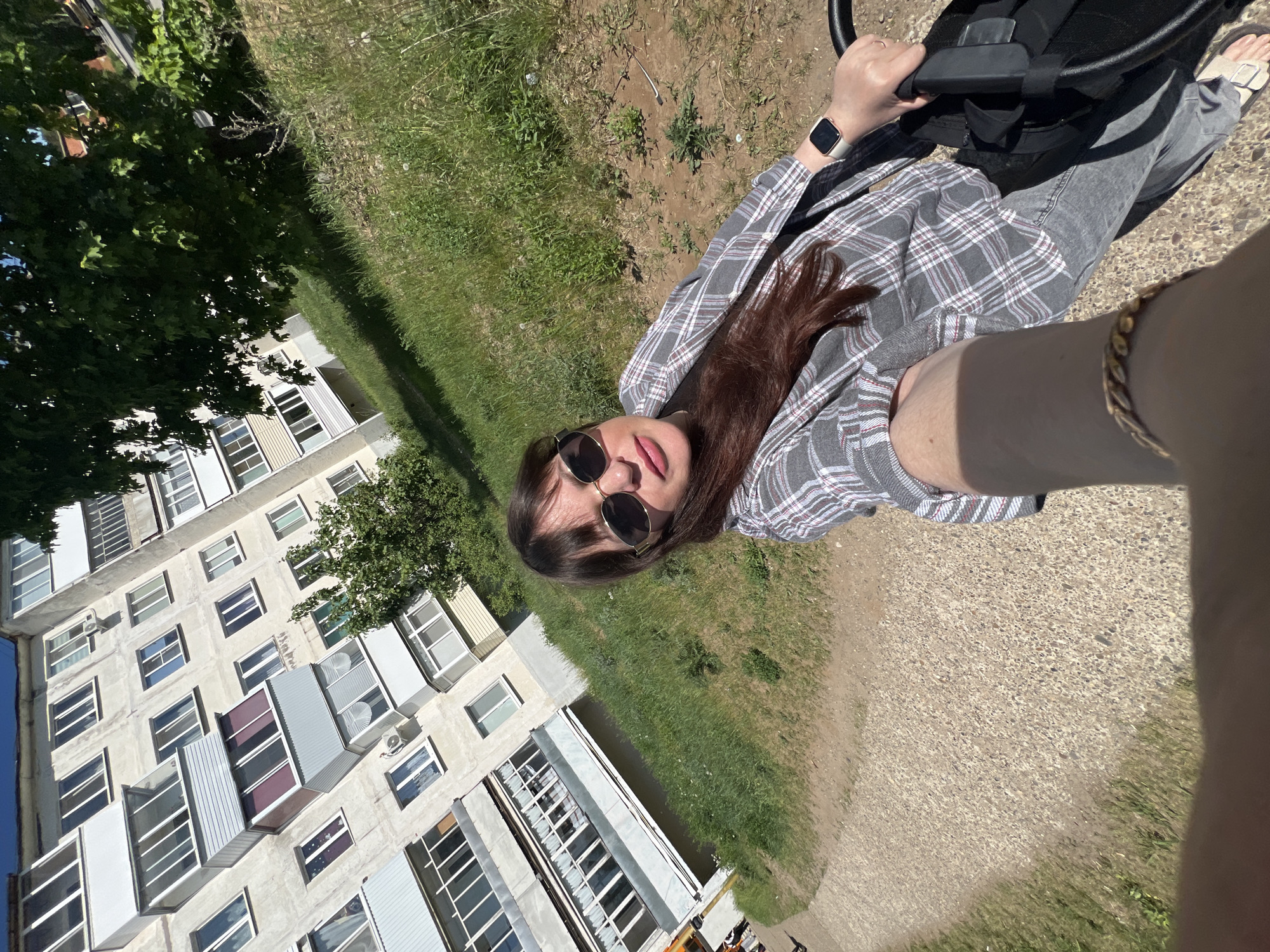 A woman wearing sunglasses and a plaid blazer is taking a selfie while holding the handle of a stroller. She is on a sidewalk next to a grassy area. In the background is a multi-story apartment building. The location is Noviy, Russia. The woman is wearing a watch. She is also wearing a bracelet. The ground is covered in dirt. The woman is also wearing gray jeans and sandals.