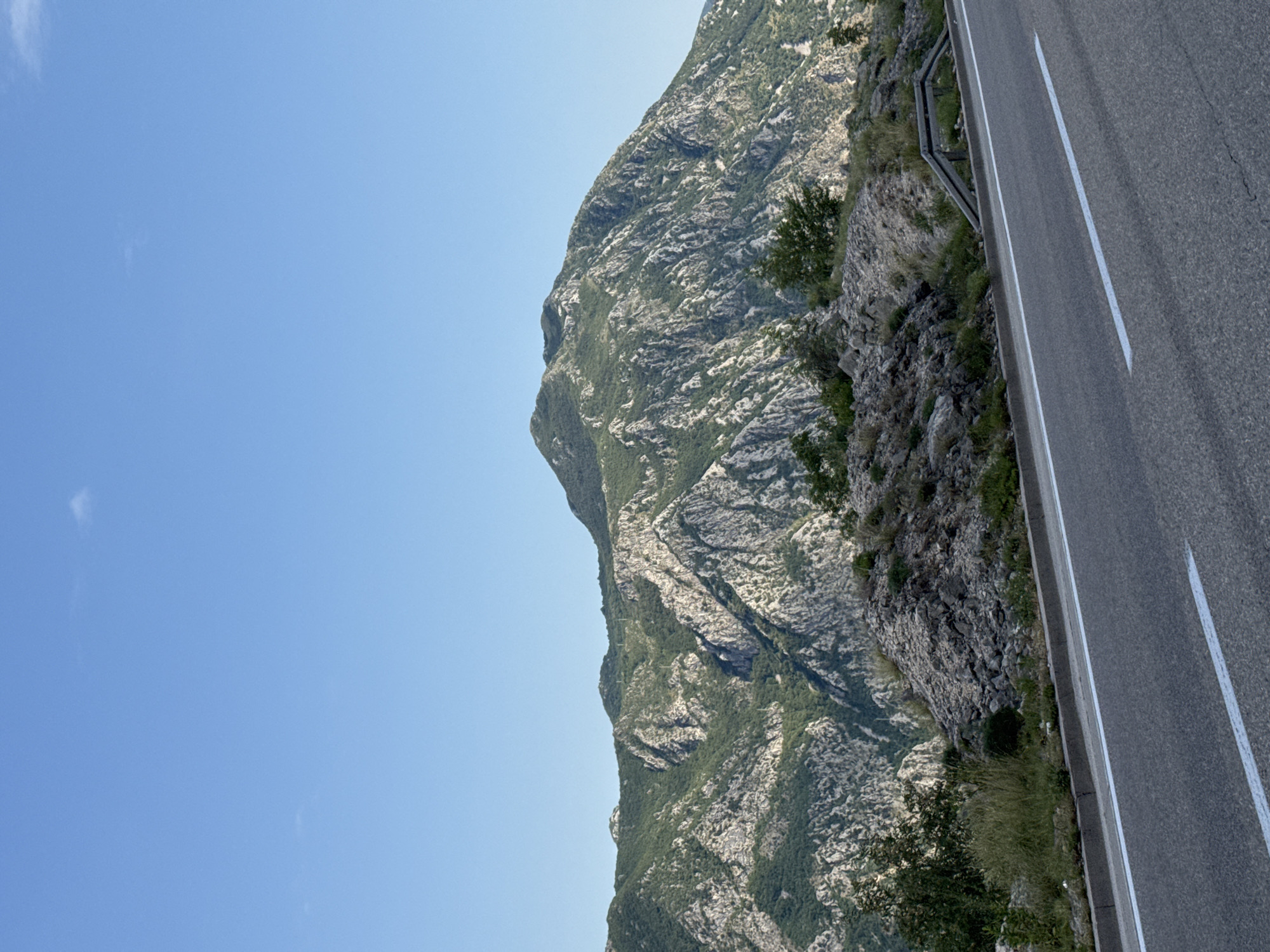 An asphalt road with white lane markings curves through the foreground, leading towards a rugged, mountainous landscape. The mountains are covered in dense green vegetation and rocky outcrops, with a clear blue sky overhead. The scene appears to be a scenic overlook or a road passing through a mountainous region near Risan, Montenegro. No people are present in the image.