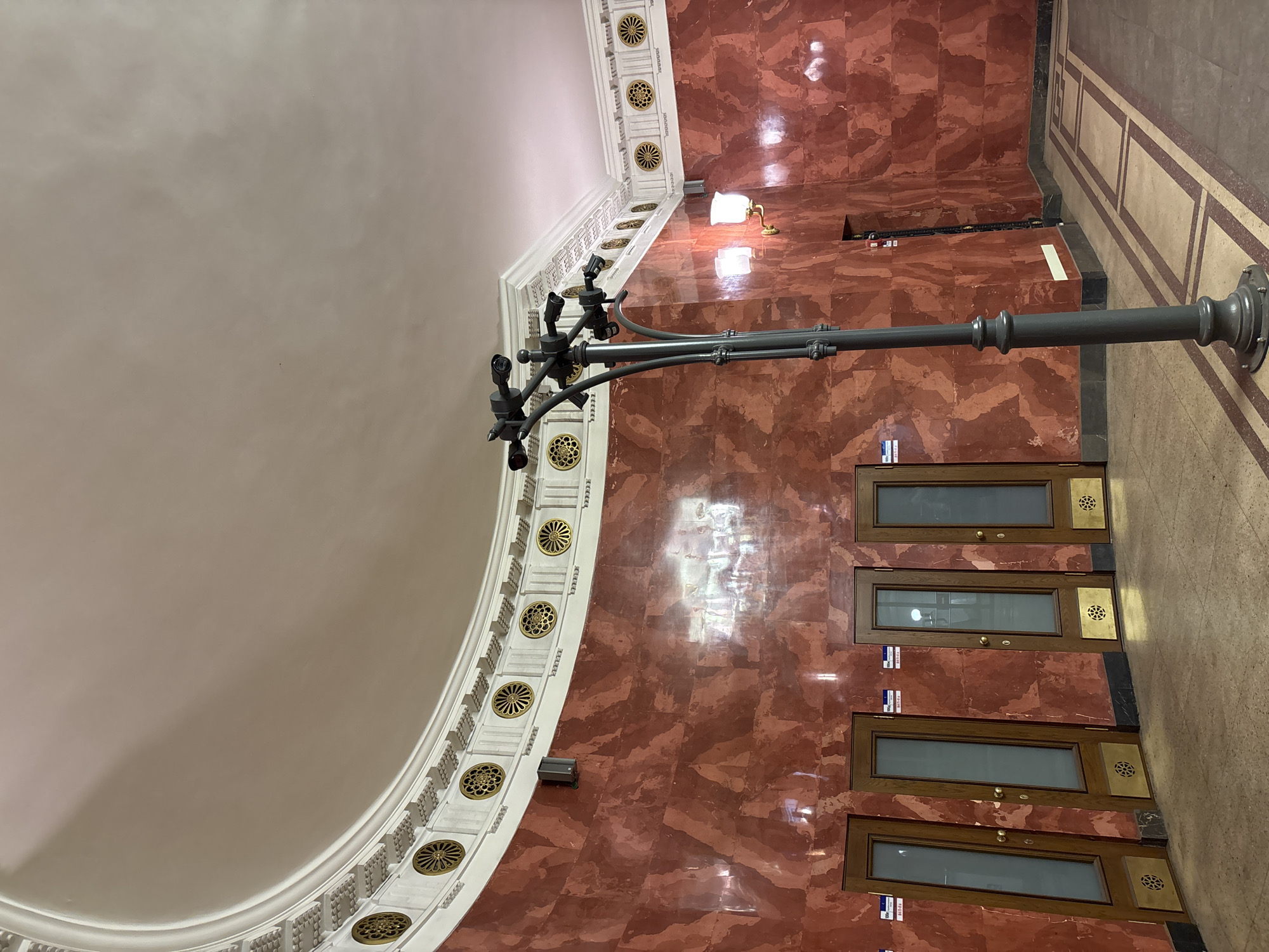 The image depicts the interior of Avtovo Metro Station in Saint Petersburg, Russia, specifically an ornate entrance hall or concourse. The scene is dominated by curved walls clad in polished reddish-brown marble exhibiting a variegated pattern of lighter red and white veining. A high, smooth, off-white curved ceiling meets the walls via an elaborate white cornice featuring repeating architectural patterns and regularly spaced round gold medallions.
The floor is tiled with light-colored, possibly beige, material, incorporating a pattern of darker rectangular outlines. In the mid-ground, a dark grey metal pole rises from the floor, branching into multiple arms near its top, each mounting a small, dark-colored cylindrical security camera; at least five cameras are visible. A single traditional-style wall-mounted lamp with a frosted glass shade and a brass base is affixed to the right wall and is illuminated.
Four identical wooden doors are arrayed along the curved left wall. Each door features a tall, narrow frosted glass panel in its upper section and a solid brass or gold-colored kickplate at the bottom, which is perforated with a circular, ornate grate design. Small white and blue labels are attached above each door frame. A black, rectangular speaker is mounted high on the left wall near the cornice. Reflections of overhead lighting fixtures are visible on the polished marble surfaces. No individuals are present in the scene, and no direct actions or interactions are depicted.