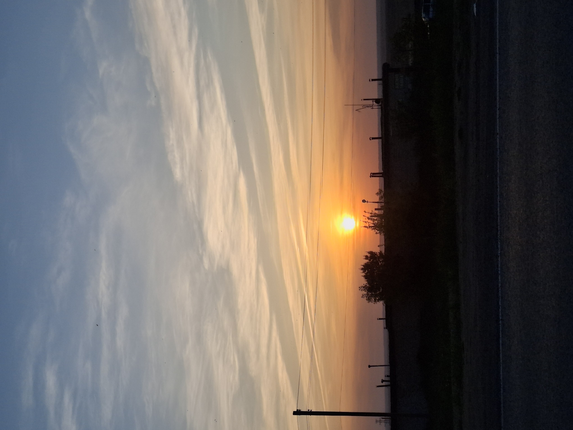 The image depicts a sunrise or sunset scene in Chaikovsky, Russia. The sun is low on the horizon, emitting a bright orange glow. The sky is filled with linear clouds, transitioning from blue on the left to orange near the sun. Power lines stretch across the horizon. A road with a white dividing line runs along the bottom right of the image. Trees and other vegetation form a dark silhouette against the orange sky. Several vertical posts are visible near the trees. The perspective of the image is horizontal, as if taken from a ground-level viewpoint.