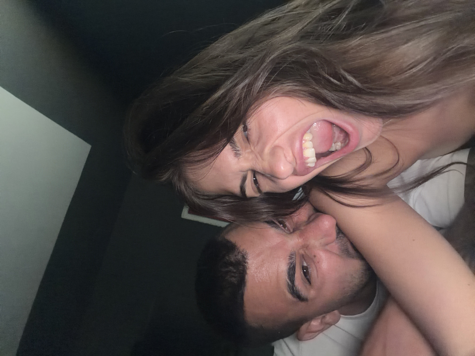 A low-angle close-up shot depicts a woman with long, brown hair, screaming widely with her mouth open, and a man with dark hair, positioned beneath her with his head turned. The woman's right arm is visible across the man's neck. Both are wearing white t-shirts. The background features a dark area on the left side of the frame and an undefined object behind the woman. The scene appears to be indoors. The location of the scene is Unknown, Unknown.
