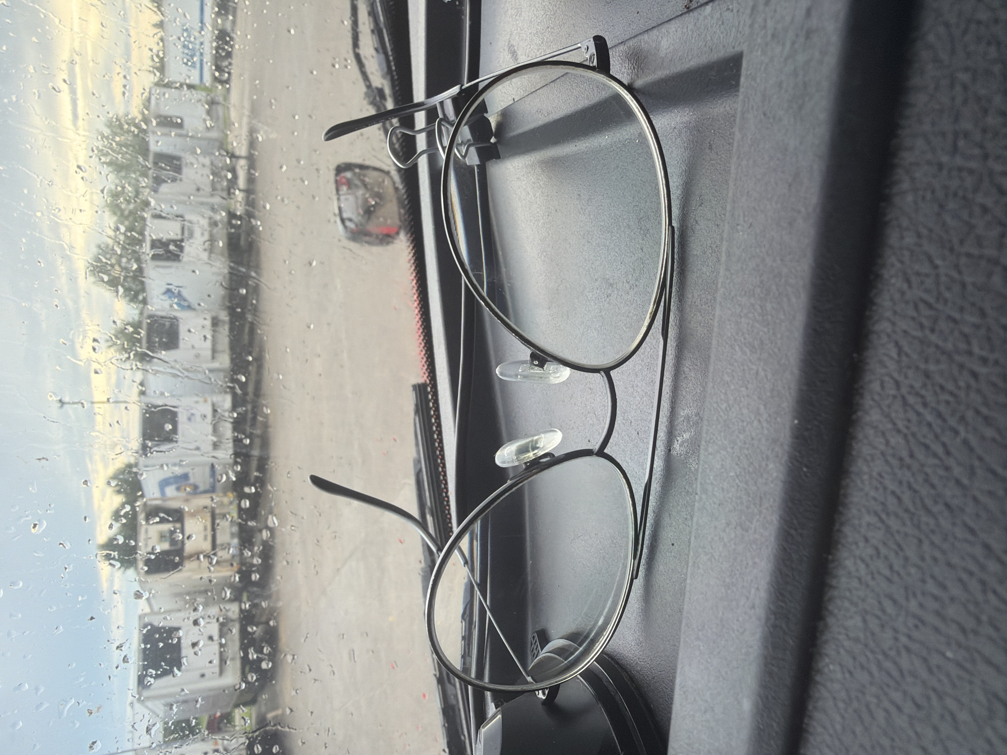 An overhead, close-up shot captures a pair of thin-framed glasses resting on a black, textured surface, possibly a dashboard, inside a vehicle. The glasses have clear, round lenses and thin silver frames. The left lens appears slightly detached or bent upwards at the frame. In the background, viewed through a rain-streaked window, a line of white refrigerated semi-trailers are parked in a lot under an overcast sky with hints of sunlight. The scene suggests a roadside stop or parking area, common in commercial trucking hubs. The weather appears to be rainy, with water droplets visible on the window. No people are visible in the image.