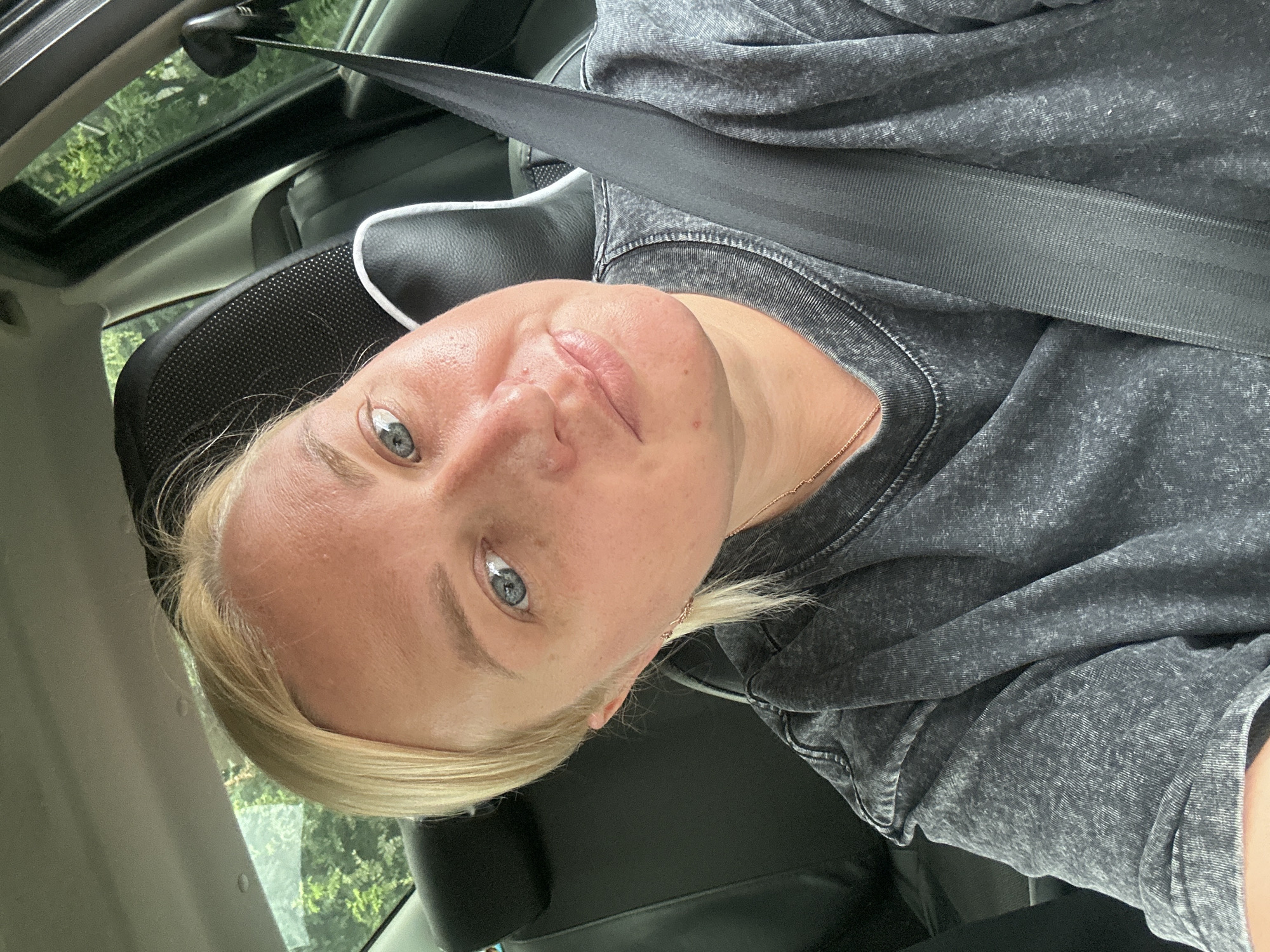 A blonde woman in her 30s with blue eyes is sitting in the driver's seat of a car, wearing a grey t-shirt and a seatbelt. She is looking at the camera with a neutral expression, her left cheek appears slightly swollen. The interior of the car is visible, with black seats and a headrest. Outside the car windows, green trees and foliage are visible, suggesting a rural or suburban setting. The image appears to be taken on a sunny day. No specific location within Chaikovsky, Russia can be determined from the visual information provided.