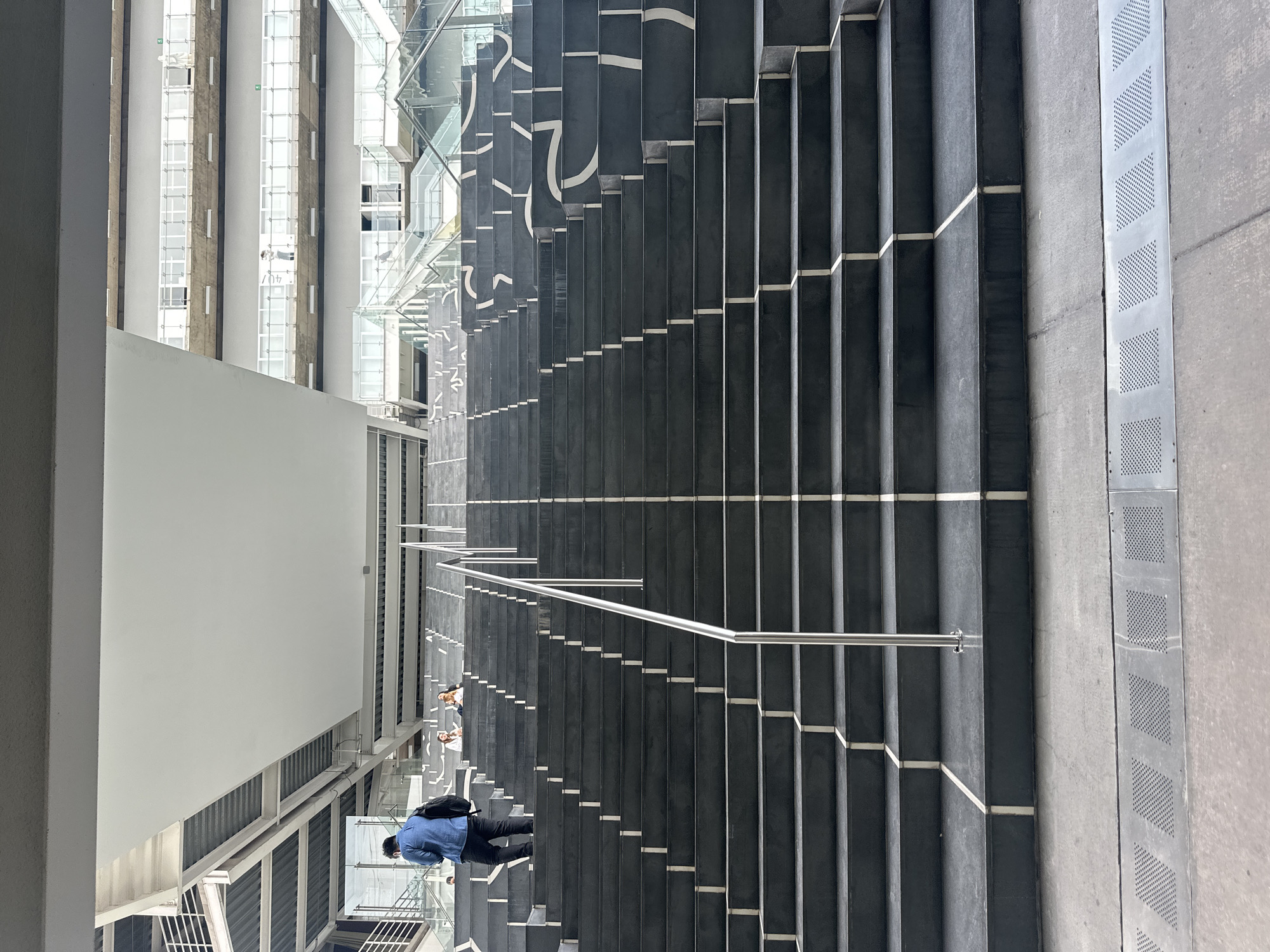 The image depicts a set of stairs with black risers and white treads, located in Mexico City, Mexico. The stairs are situated inside a modern building with glass and metal architecture. A metal handrail runs alongside the stairs. A man in a blue shirt and black pants is walking down the stairs, along with two other people ahead of him. A patterned metal grating borders the stairs on one side. Adjacent to the stairs is a tall building facade with visible windows.