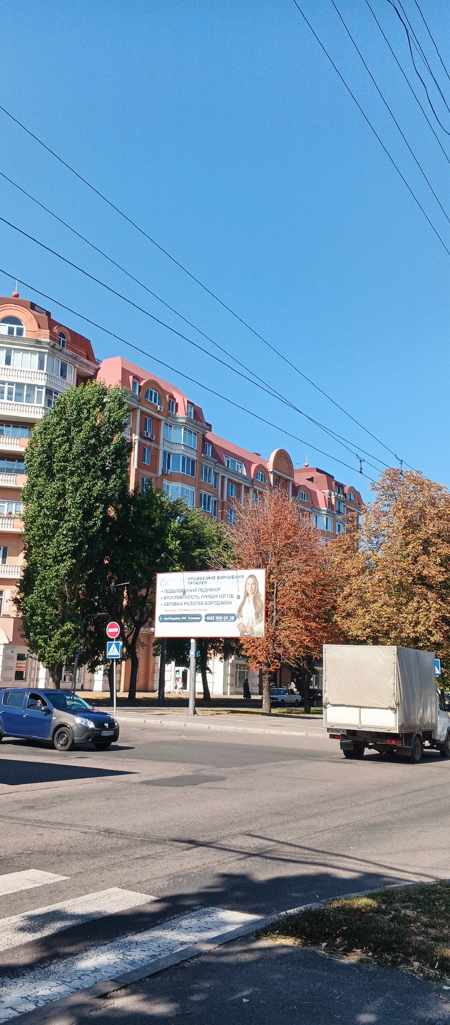 The image shows a street scene in Cherkasy, Ukraine. A blue and gray car is driving on the road, and a white truck with a covered bed is parked on the side. A billboard is visible in the background, displaying an advertisement for professional problem-solving services, including podiatry and pedicure. The billboard also features a picture of a woman. The buildings in the background are modern apartments with red roofs. The sky is clear and blue, and the trees have autumn foliage. There is a pedestrian crossing on the road in the foreground.