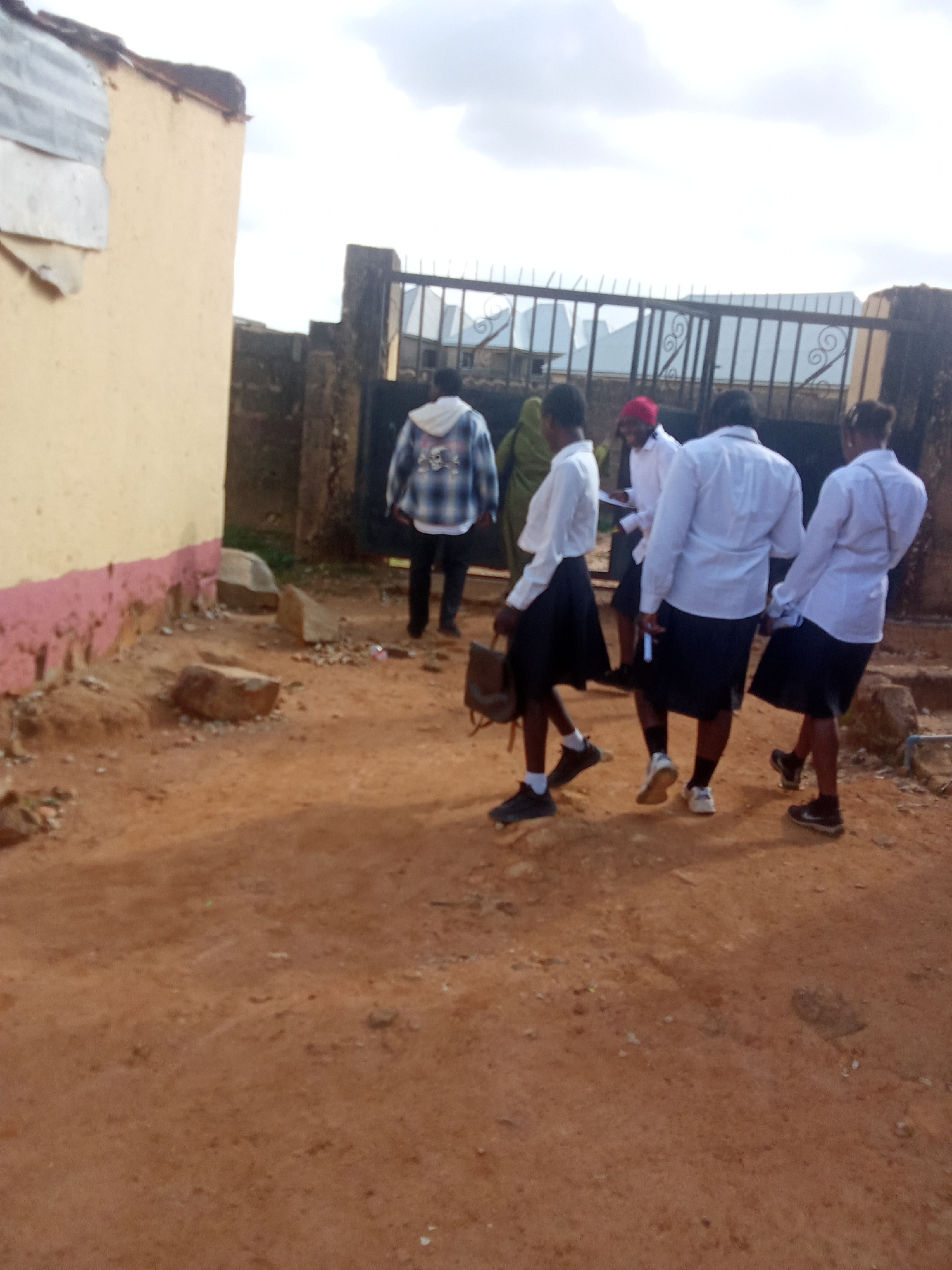 In Barakin Akawo, Nigeria, a group of individuals walks near a textured, light-colored wall with a pink-and-red painted base. A dark metal gate with ornate scrollwork stands behind them, with a building visible through it. The ground is composed of reddish-brown dirt with scattered rocks and debris. One person in the foreground wears a checkered jacket over a white shirt, black pants, and dark shoes. Behind them, three individuals in white shirts and black skirts walk away from the camera. One of these individuals carries a brown bag, and another wears a red headscarf. The sky is overcast with clouds.