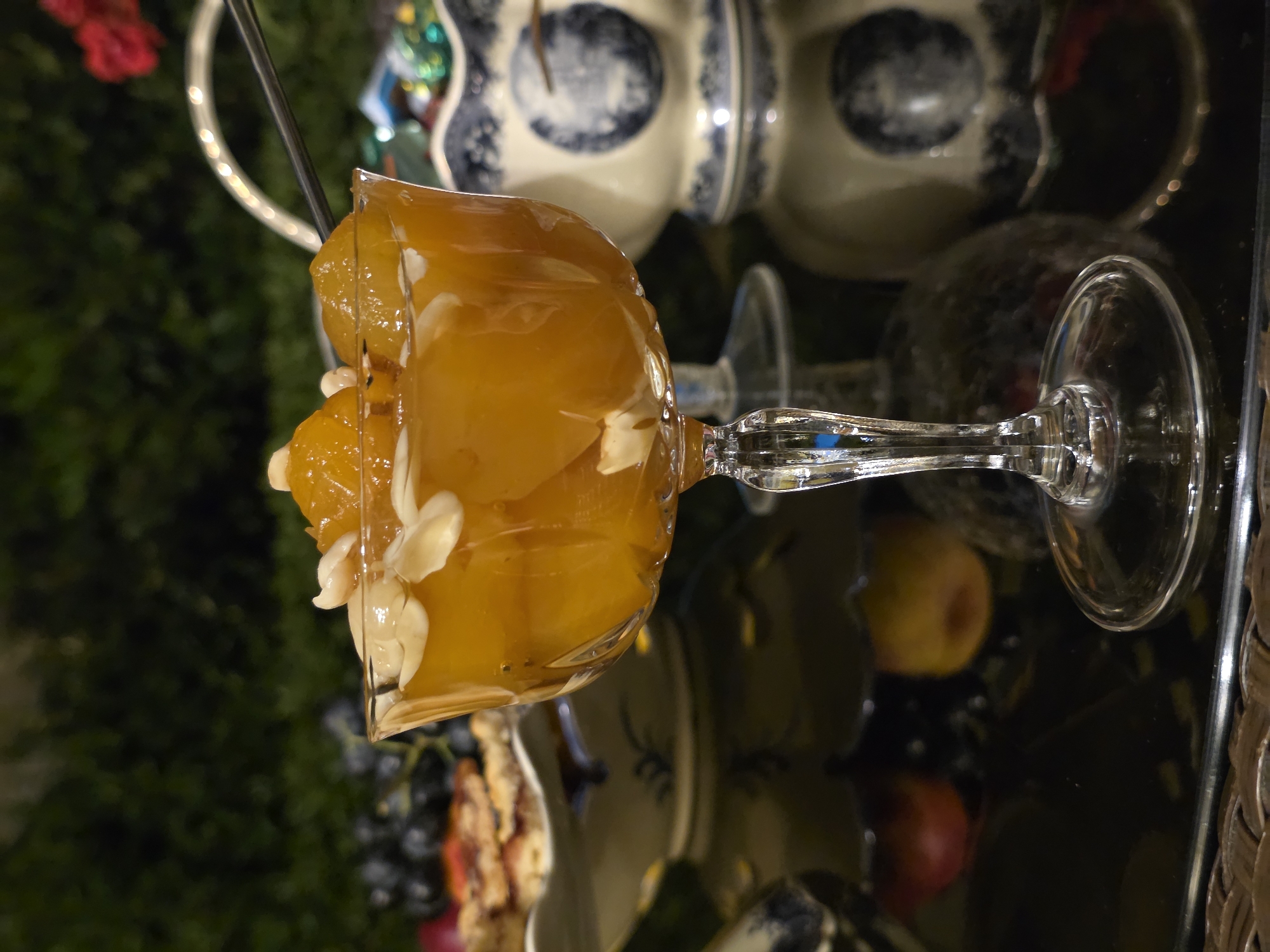 A close-up, low-angle shot captures a glass coupe filled with preserved apricots and slivered almonds. The coupe, made of clear, faceted glass, rests on a reflective black surface, possibly a table. The apricots are a vibrant orange hue, submerged in a light syrup. In the background, a dark green hedge serves as a backdrop. To the left, a portion of a platter with a patterned edge is visible, holding what appears to be food items, including dark grapes and a sliced pastry. To the right, a blue and white porcelain pitcher with intricate designs sits on the reflective surface. A silver spoon is partially visible, extending from the coupe towards the pitcher. The lighting suggests an outdoor evening setting, with subtle reflections on the table and a warm, ambient glow. The city of Buzovna, Azerbaijan, is not visible in this image, nor are there any people or discernible actions or interactions. The scene is static and focuses on the dessert.